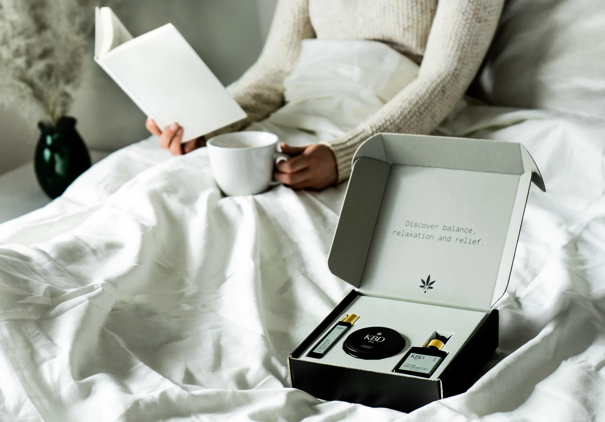 Person sitting on bed with a mug, reading a book, and a CBD product box open in front of them.