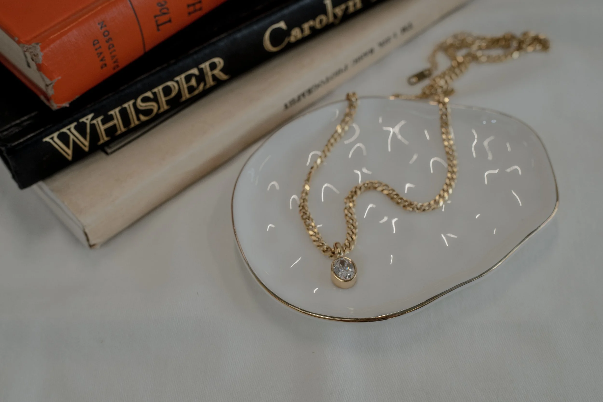 A gold necklace with a single round jewelry pendant rests on a white decorative dish with gold edges, placed on a white surface, with a stack of books partially visible in the background.