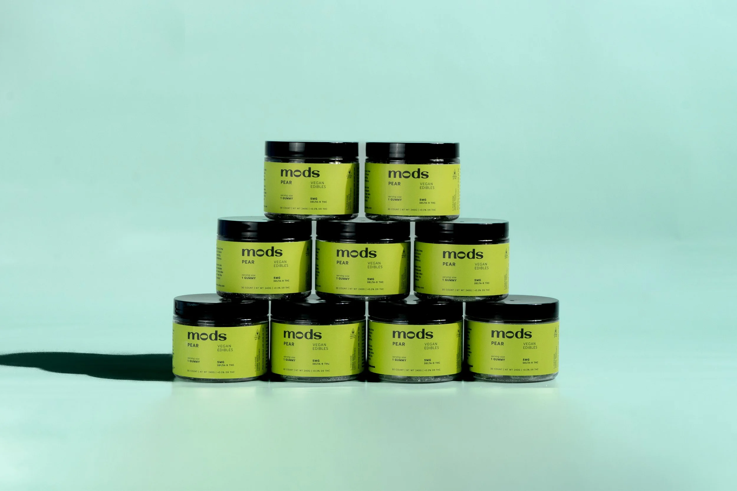 Stack of nine green containers labeled "mods" with flavor "pear" and indicating vegan edibles containing 5 mg of THC.