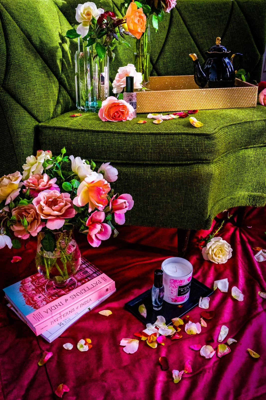 A green velvet armchair decorated with flowers, including roses in vases on top and roses scattered on a pink cloth and floor nearby. Items like a tea kettle, nail polish, candles, and books are also on the chair and floor.