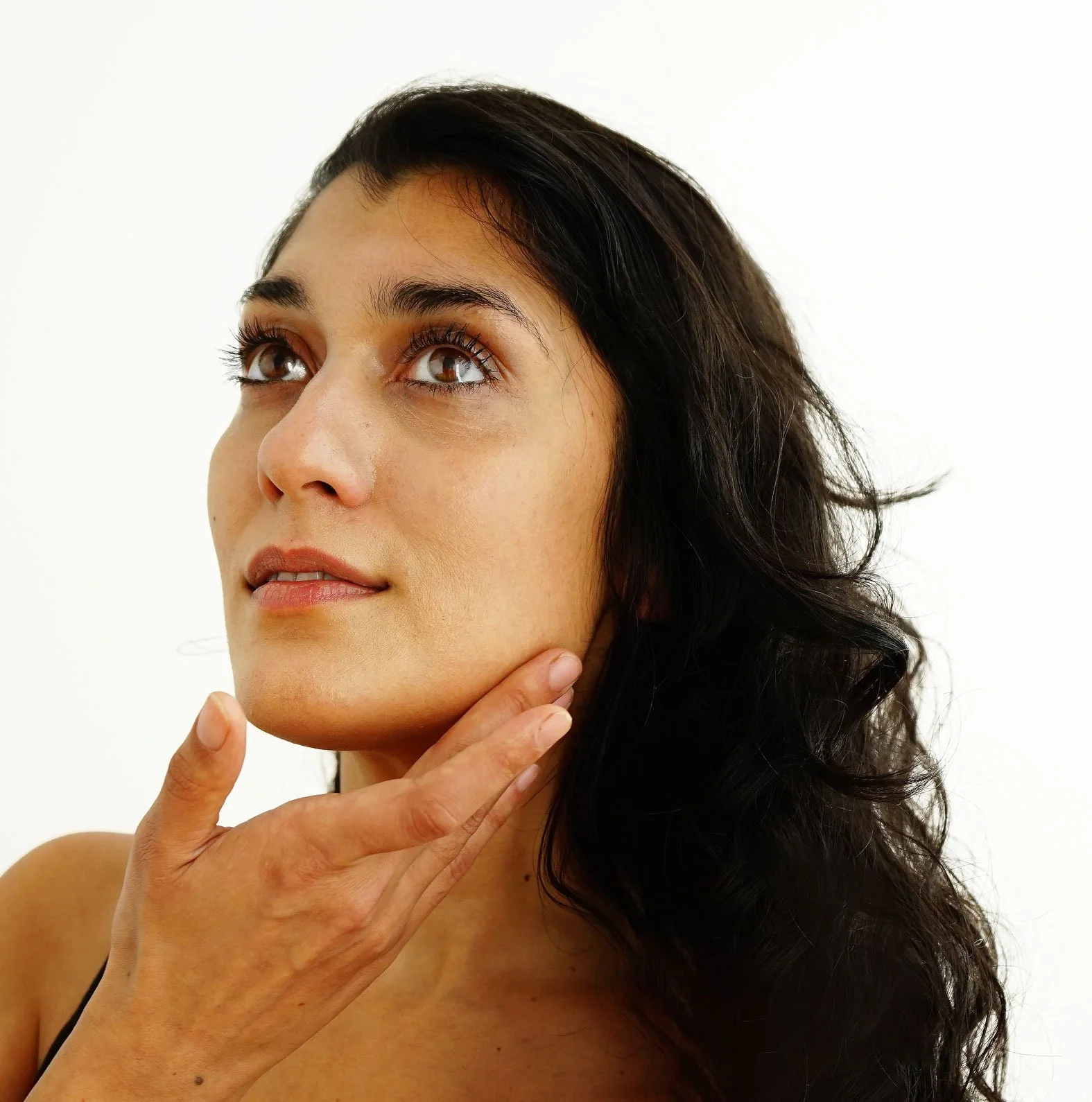 Close-up of a woman with dark, wavy hair, looking upward, touching her jawline with her fingers, against a plain white background.