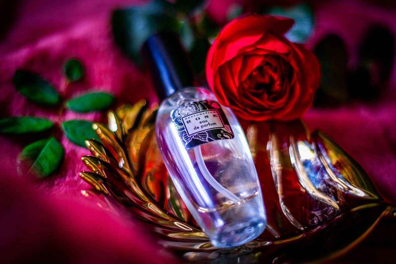 A clear perfume bottle with a black cap, labeled 'TROISIEME' and 'eau de parfum,' resting on a reflective gold tray. A red rose and green leaves are nearby, set on a pink textured fabric background.