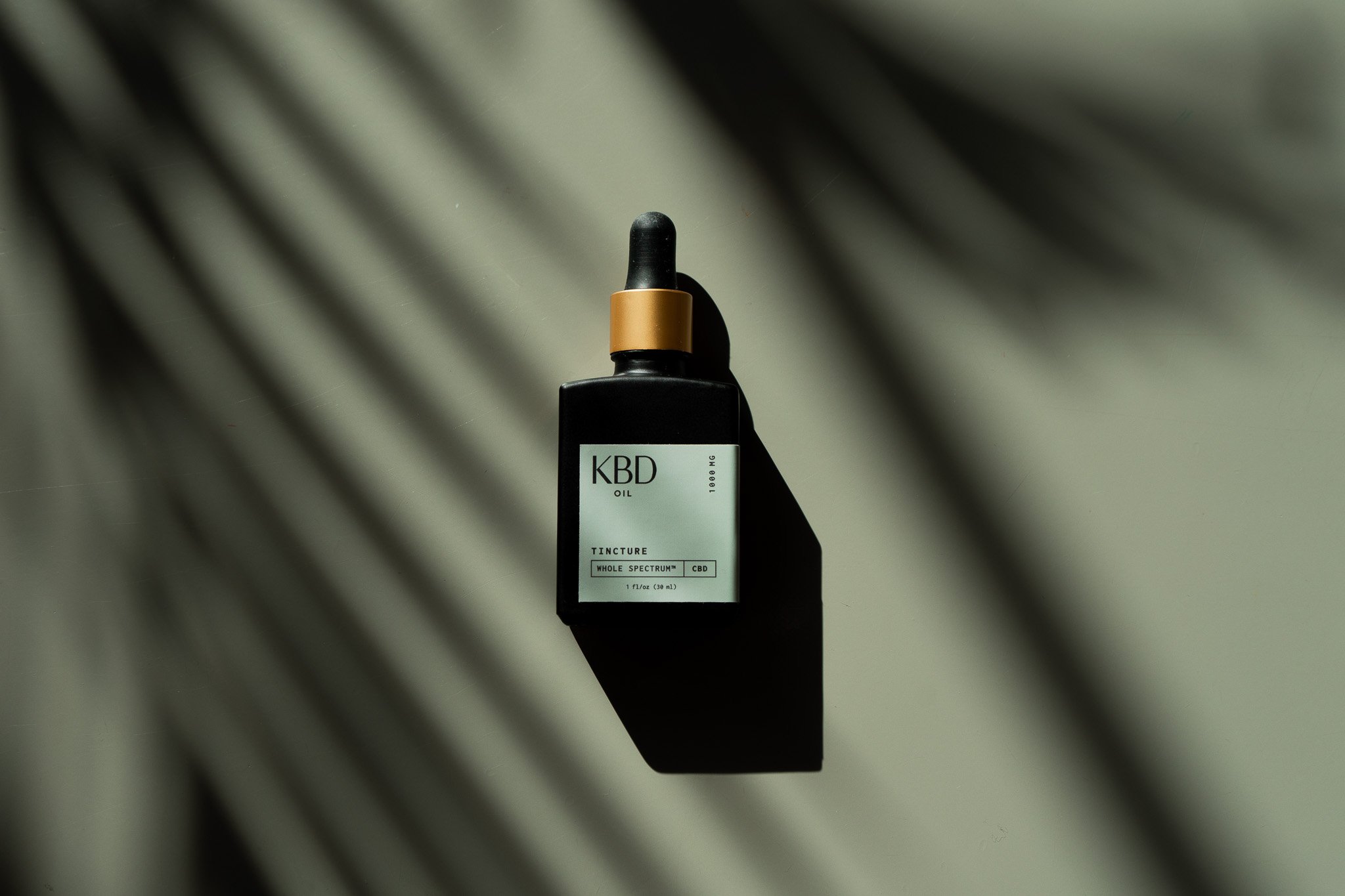 A black bottle with a dropper cap, labeled 'KBD OIL' and 'TINCTURE WHOLE SPECTRUM CBD', resting on a light surface with shadows cast by nearby objects.