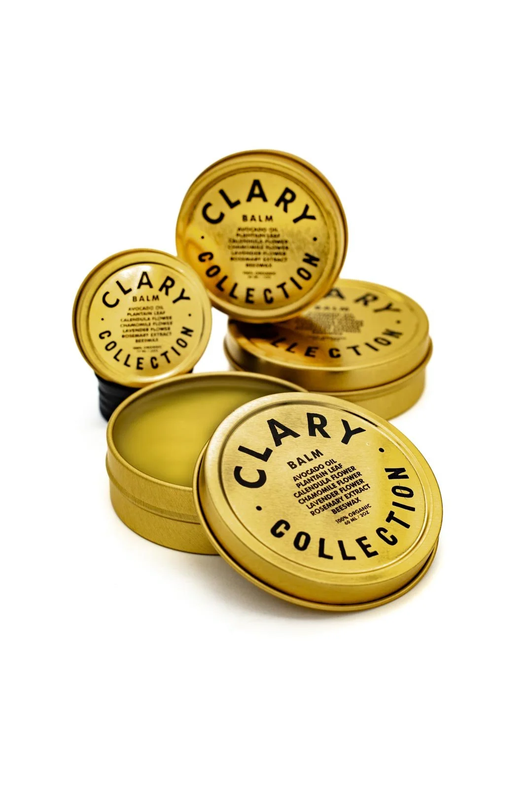 Gold-colored round tins labeled 'Clary Collection' with one open showing yellow balm inside. The labels describe the balm's ingredients: avocado oil, plantain leaf, calendula, chamomile, lavender flower, rosemary extract, beeswax.