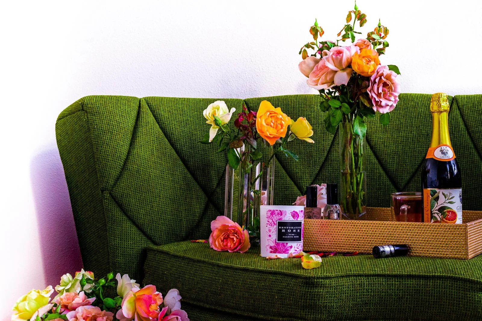Green velvet sofa adorned with assorted pink, white, yellow, and orange roses in glass vases, with perfume bottles and a box on a tray, and a bottle of sparkling wine to the right.