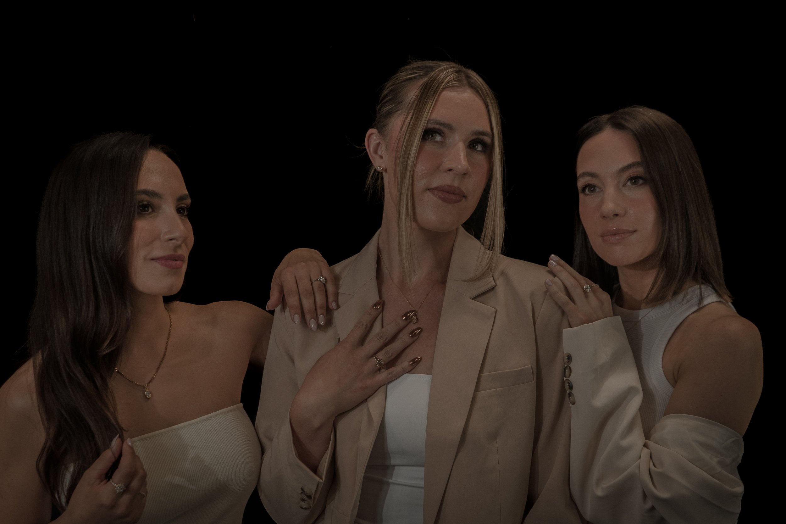 Three women pose together against a black background, dressed in light-colored outfits with jewelry, displaying confident expressions.