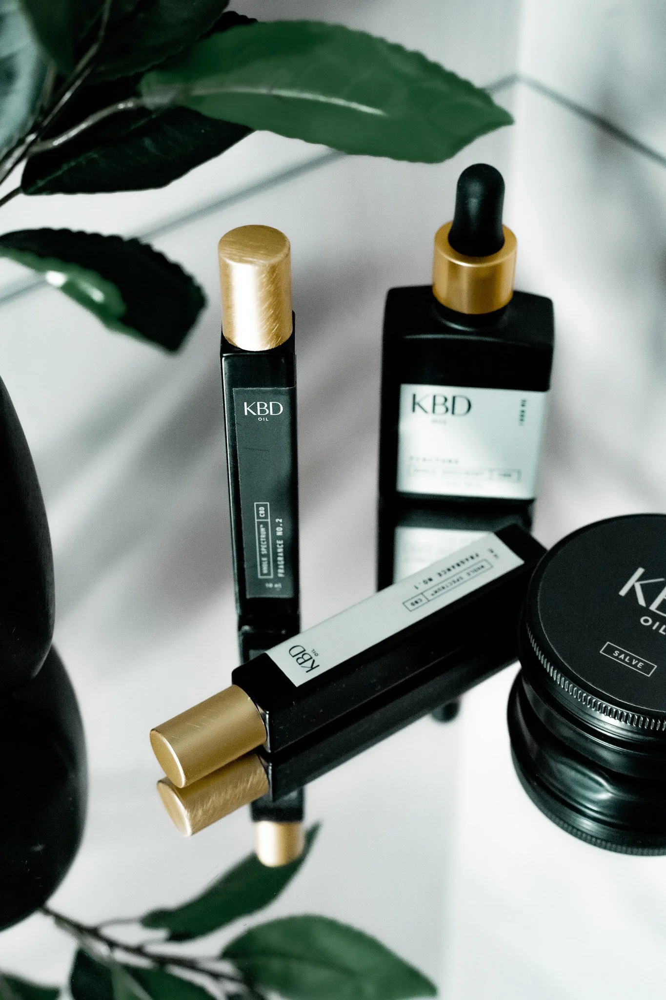 Collection of KBD skincare and cosmetic products, including a dropper bottle, matte black tubes, and a jar, placed on a reflective surface with green leaves in the background.