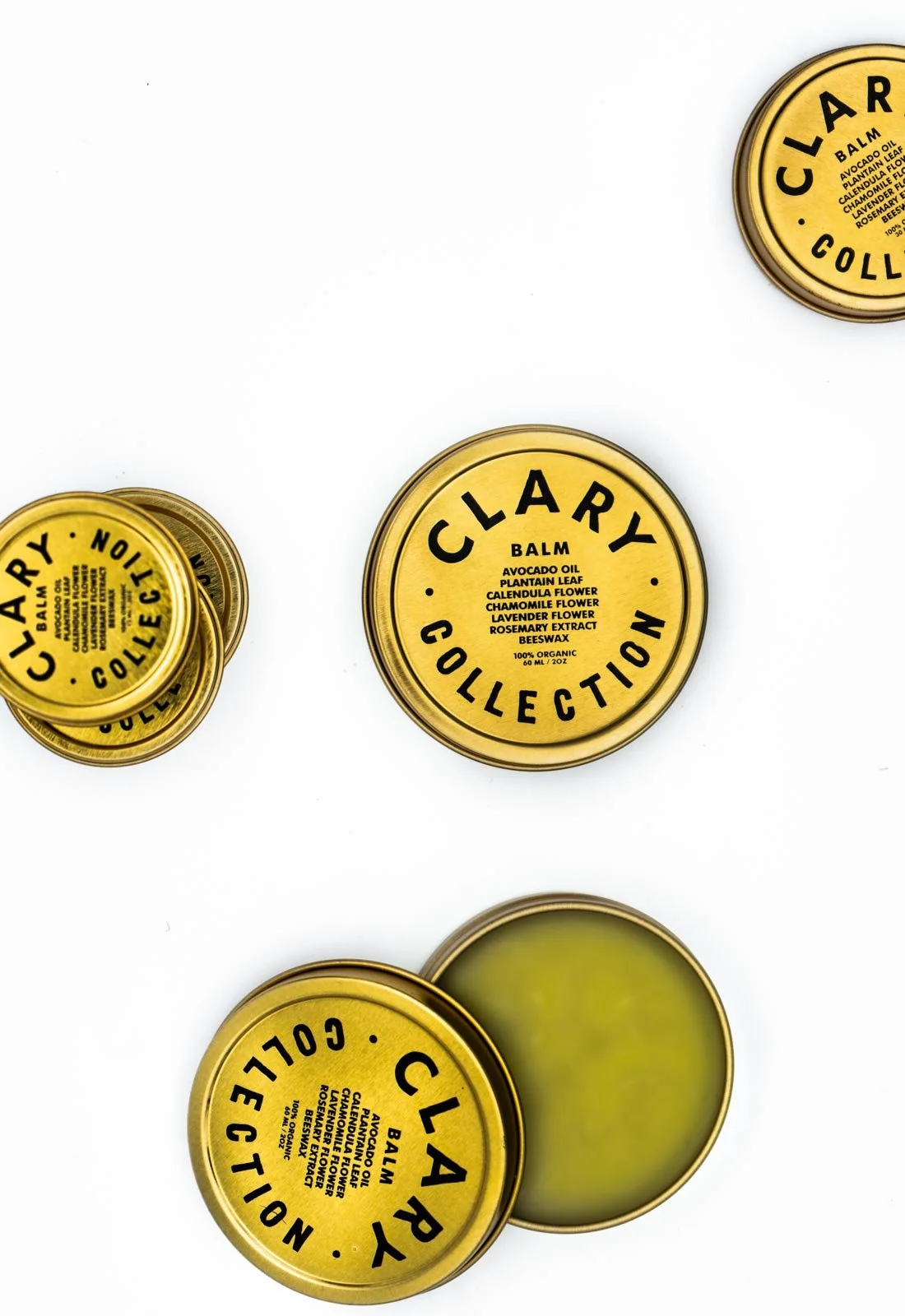 Top view of multiple yellow tins of Clarcy collection balm with ingredients like avocado oil, calendula flower, chamomile flower, lavender flower, rosemary extract, and beeswax, some open showing the balm inside, on a white background.