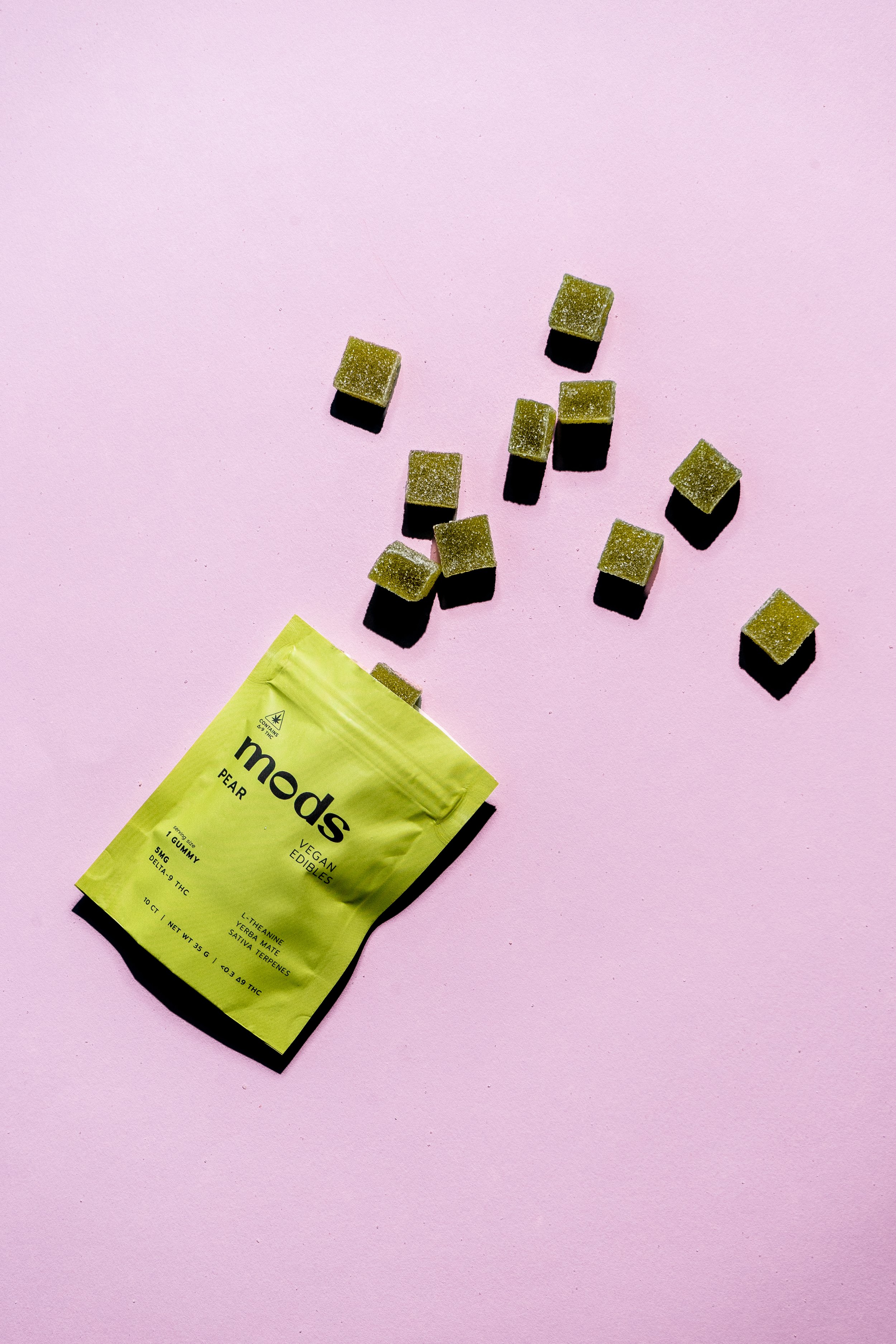Green packet of 'mods' vegan edible gummies labeled 'Pear,' with several green square gummies scattered on a pink background.