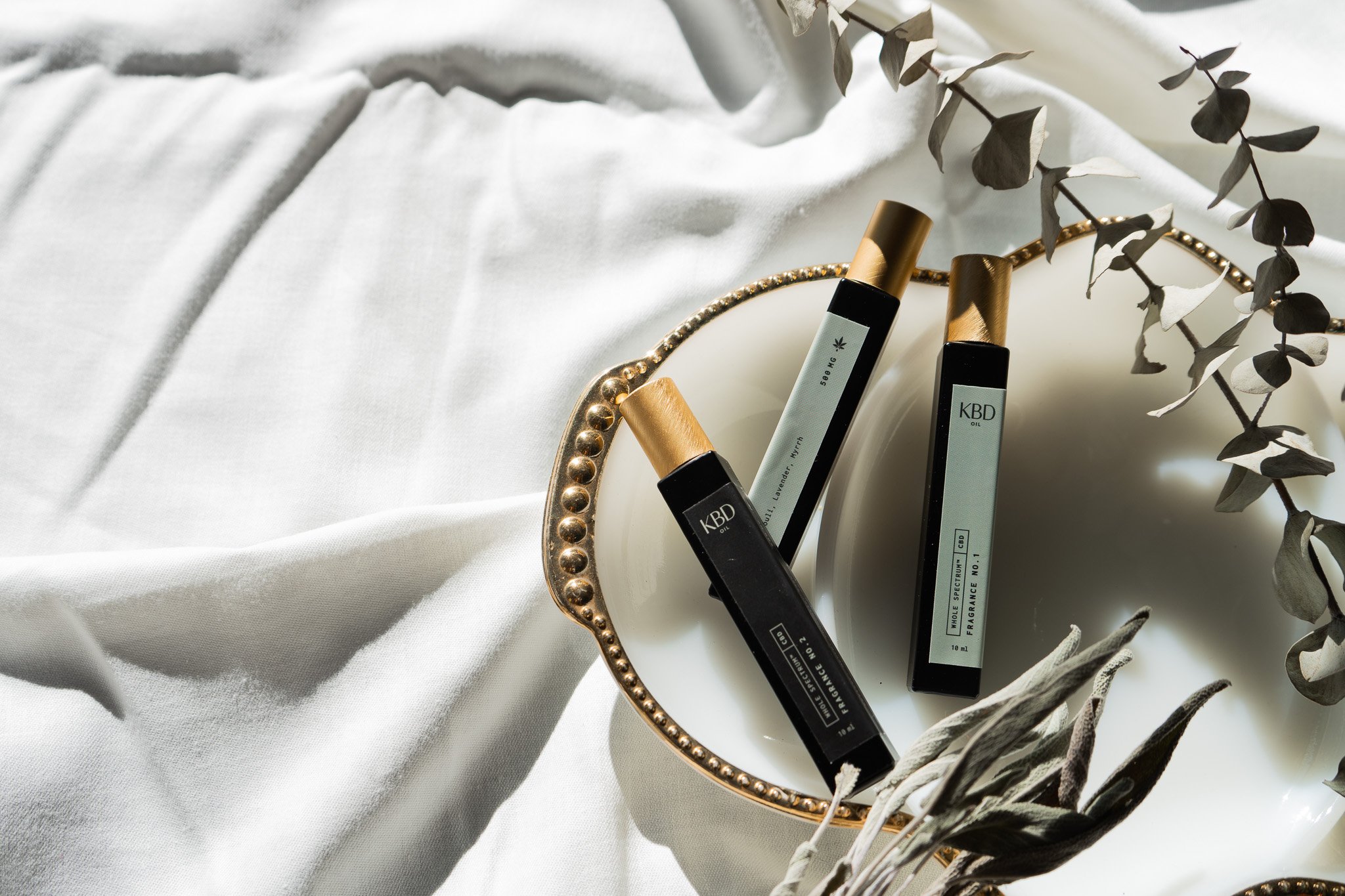 Three CBD oil bottles with black bodies and gold caps placed on a white ceramic dish with a gold rim, surrounded by white fabric and dried eucalyptus leaves.