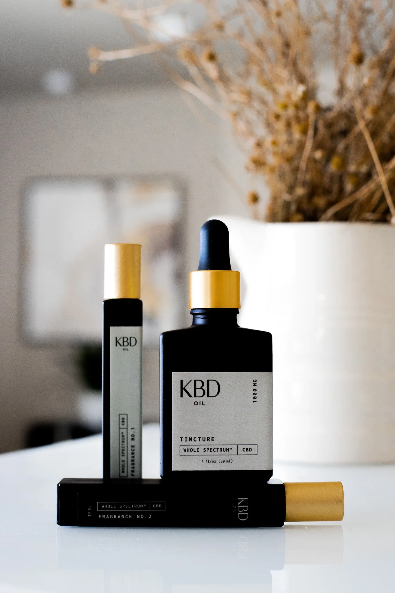 Three black bottles of CBD oil products with gold caps on a white surface, with a background of dried flowers in a white vase.