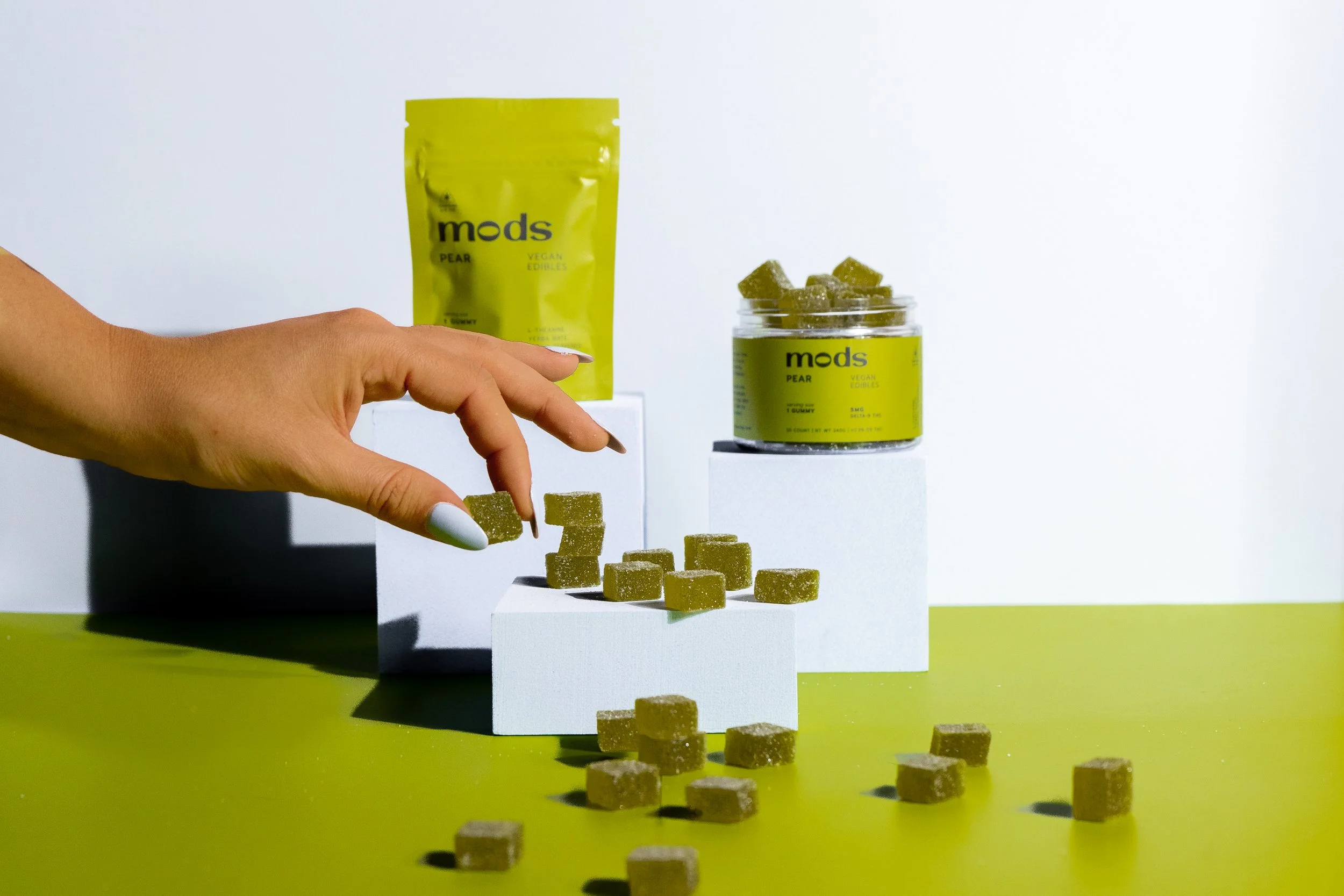 A hand with painted nails reaching to pick up a green pear-flavored gummy from a display of other gummies. In the background, there is a green bag and a jar of pear-flavored gummies labeled 'mods.'