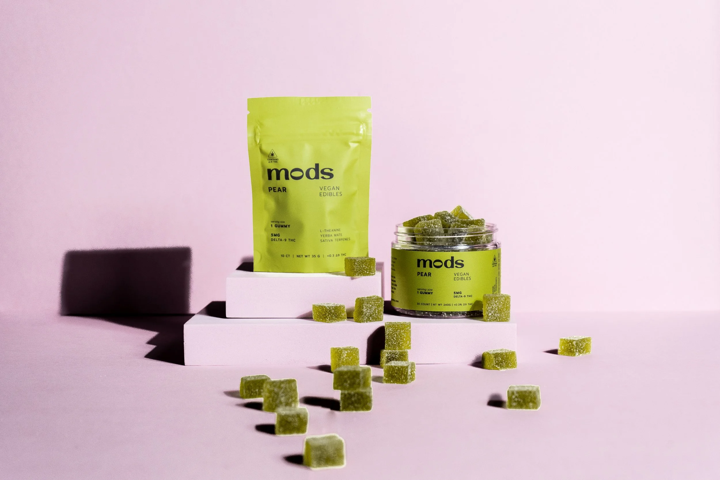 Green packaging of Mods Pear vegan edibles with a jar of gummy candies and scattered gummies on a pink background.