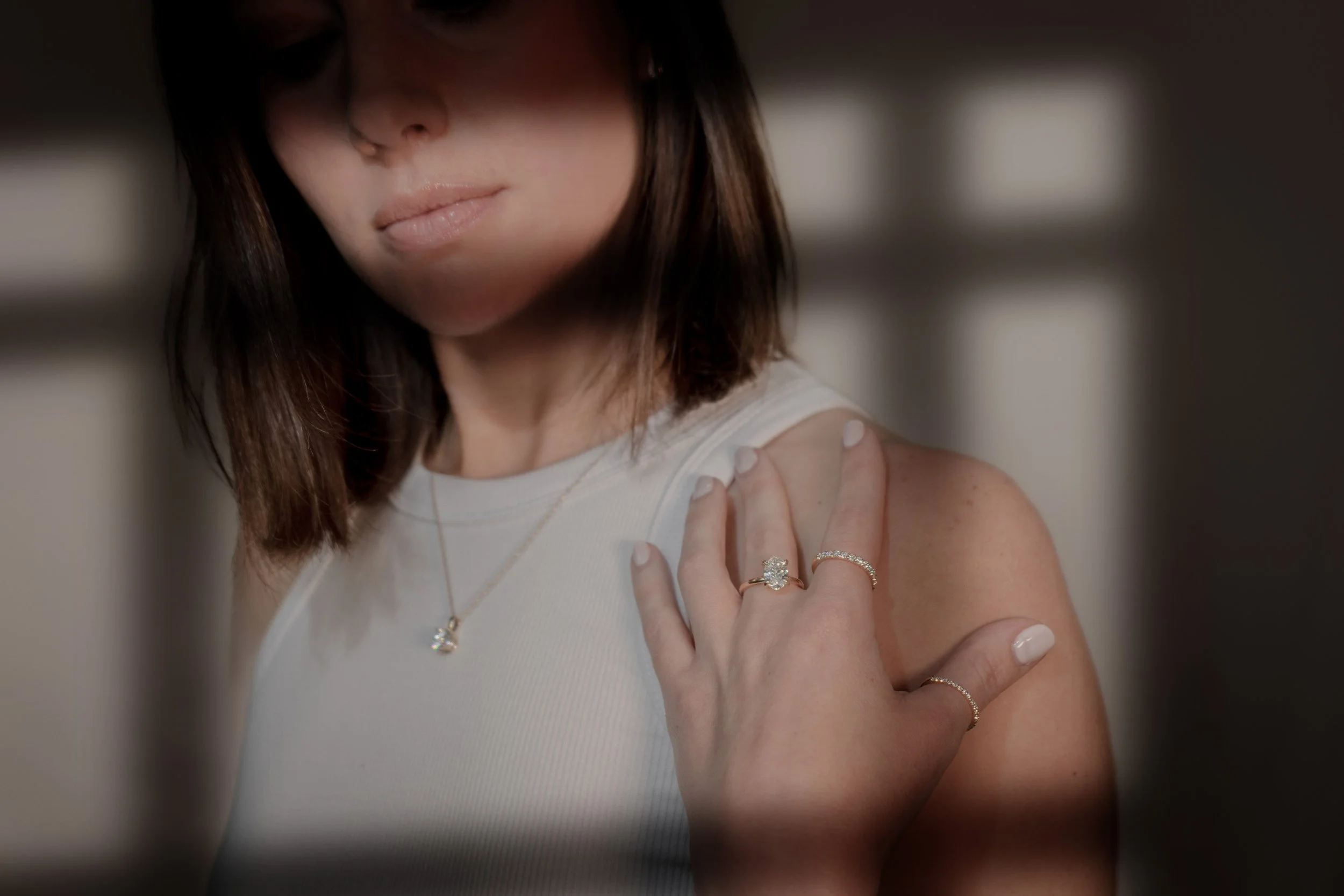 A woman with dark hair and light skin showing her shoulder. She is wearing a white sleeveless top, a gold necklace with a star-shaped pendant, and a gold ring with a large gemstone on her finger. Her nails are painted white.