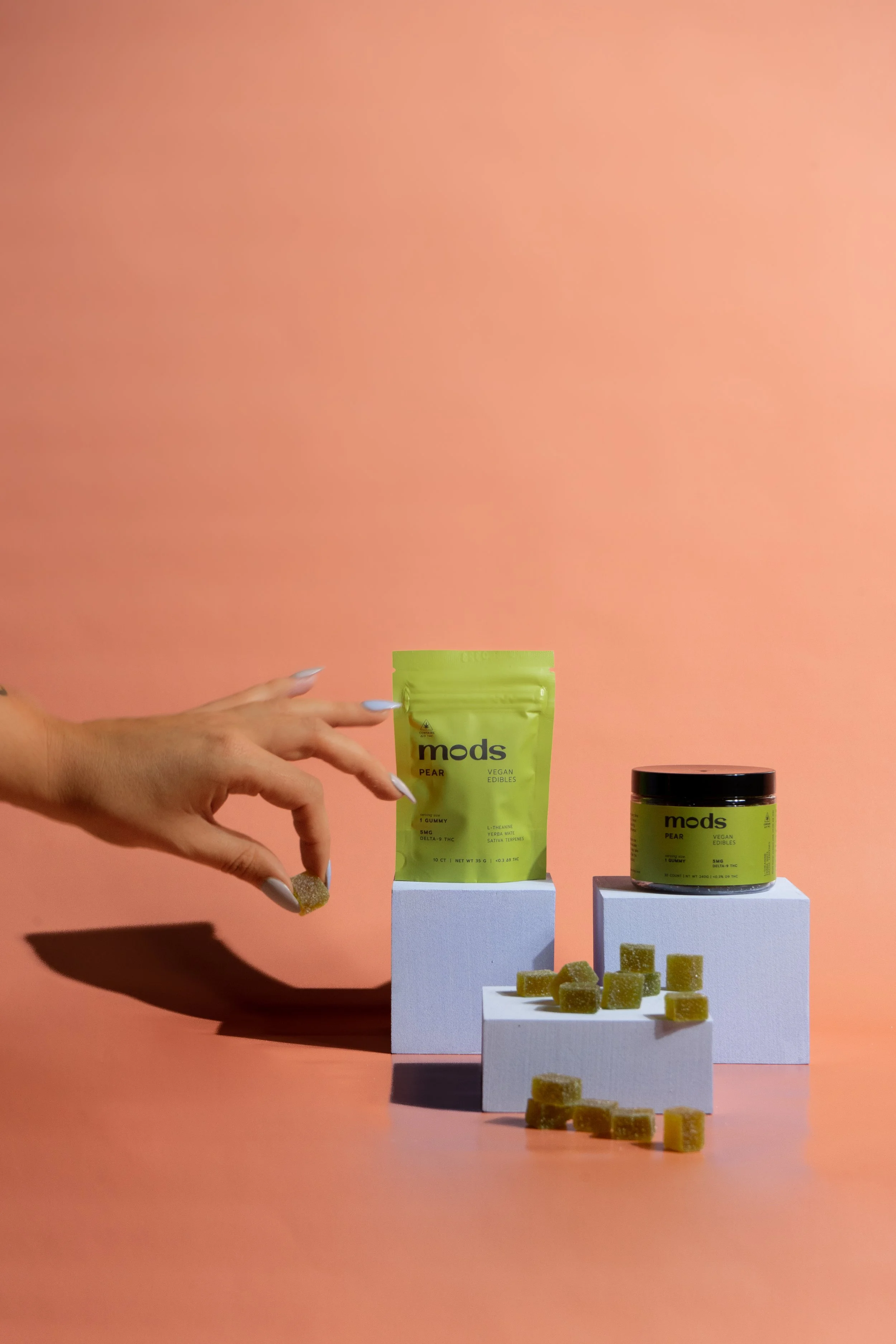 A hand holding a green gummy candy with the label 'mods' and 'PEAR VEGETAL EDIBLES' is seen against a pink background. The gummy is near two products: a green bag labeled 'mods PEAR' and a green jar with the same label, both placed on white platforms