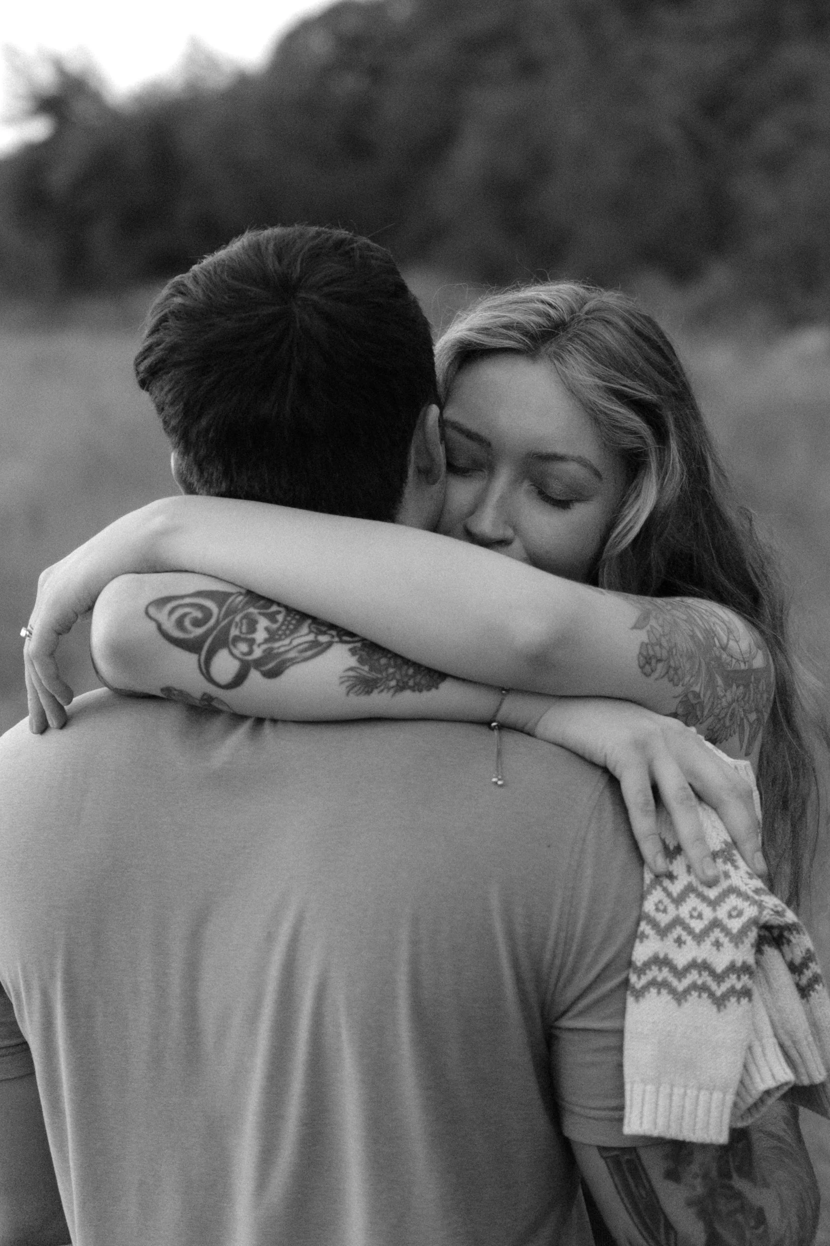 A black and white photo of a couple hugging outdoors, with the woman’s eyes closed and embracing the man.