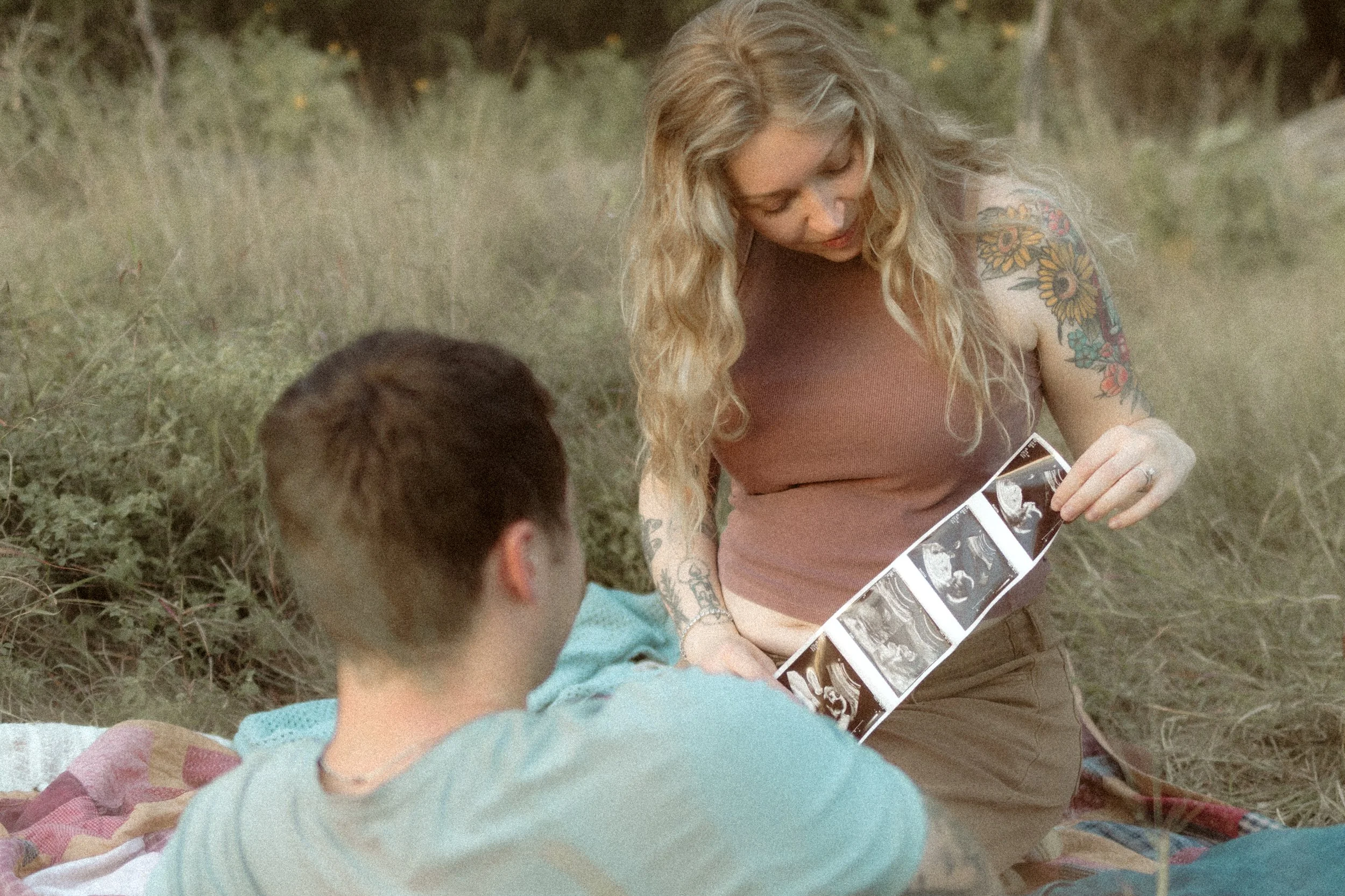 A woman with long blonde hair and tattoos on her arms, wearing a sleeveless top, is sitting outdoors on a blanket, looking at ultrasound photos. A person with short dark hair, wearing a light-colored shirt, is sitting in front of her, facing away fro