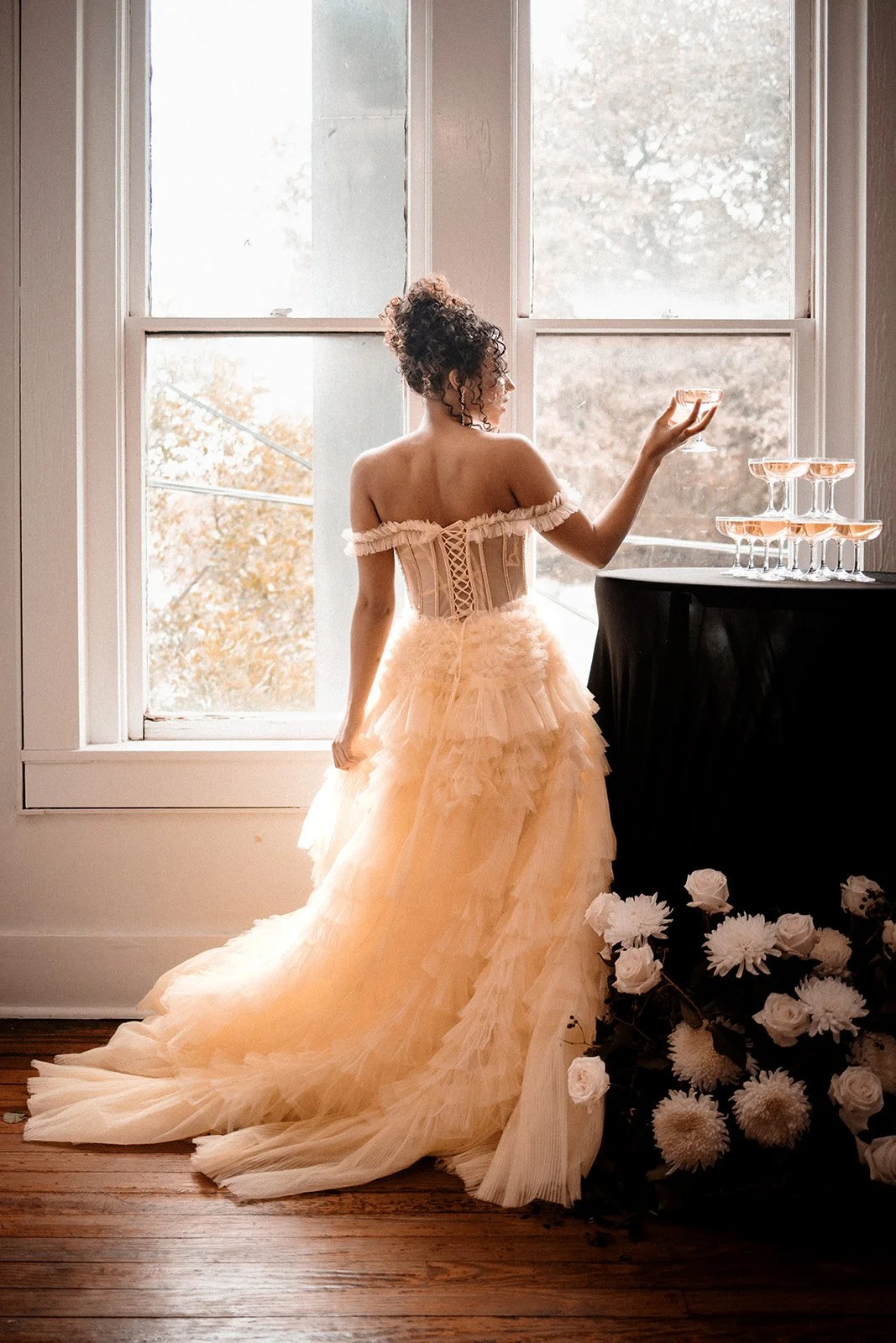 A woman in a beige, ruffled gown with an off-the-shoulder corset bodice stands by a large window, holding a glass of champagne, with a tier pyramid of champagne glasses on a table beside her and a bouquet of white flowers on the floor.
