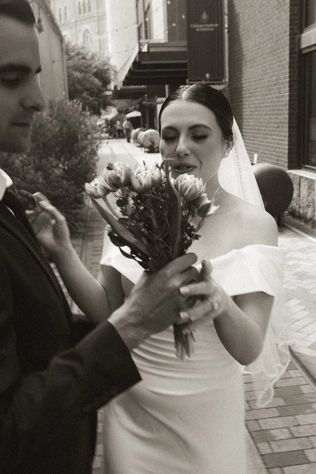 Black and white photo of a bride holding a bouquet of flowers while a person adjusts her dress or bouquet.