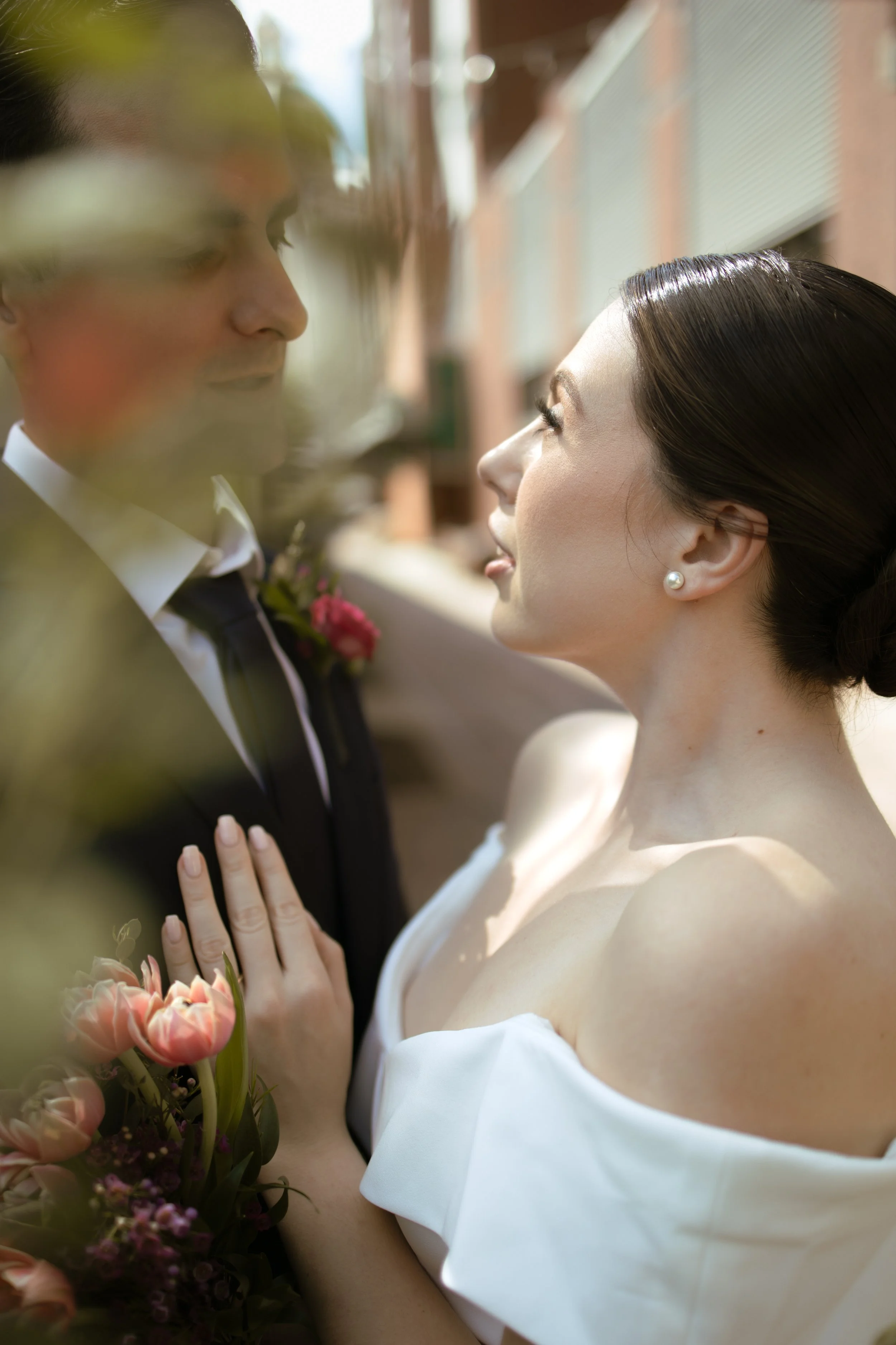 A bride and groom looking at each other closely outdoors on their wedding day, with the bride holding a bouquet of pink flowers.