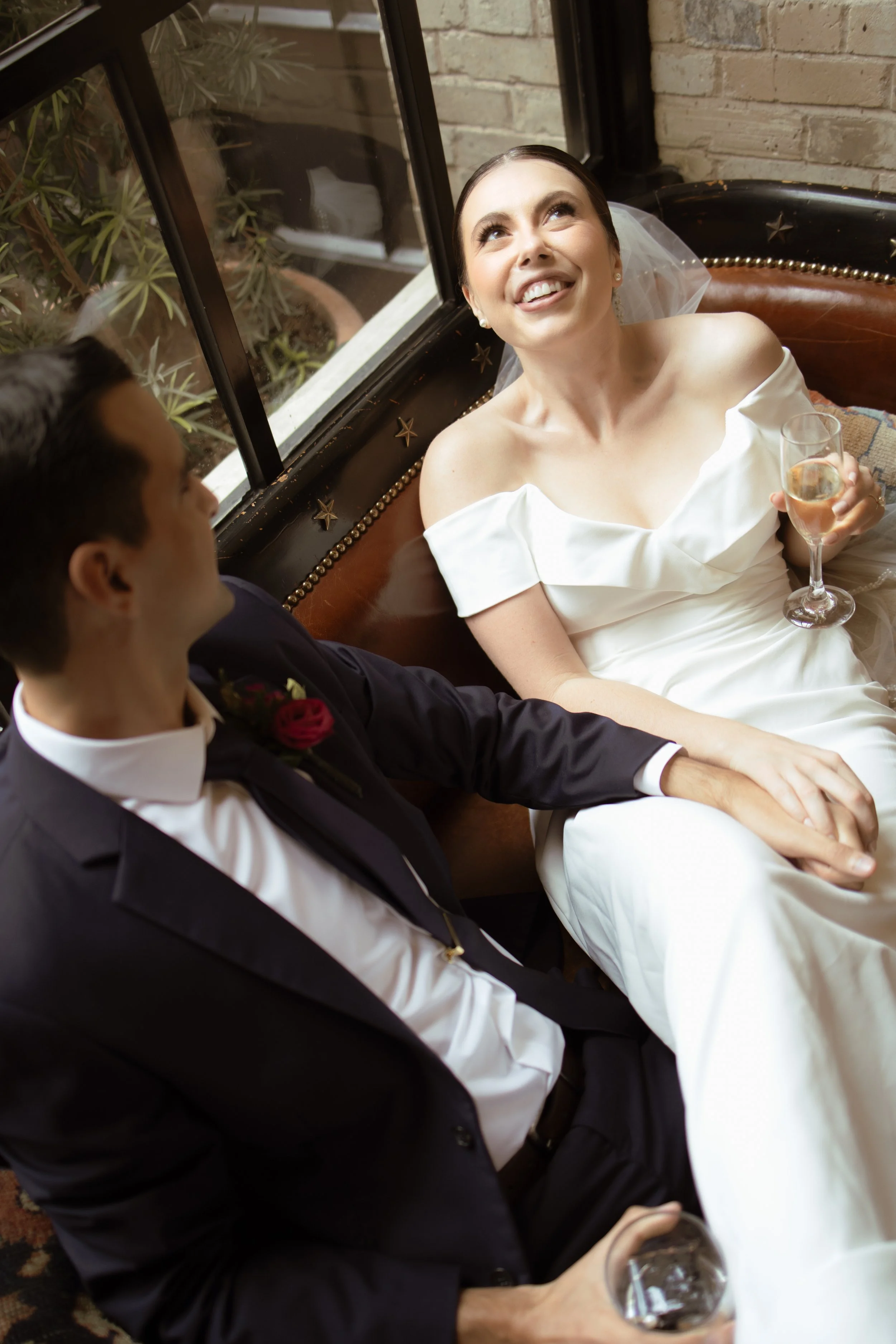 A bride and groom sitting on a couch, holding hands and smiling, with the bride holding a glass of champagne.