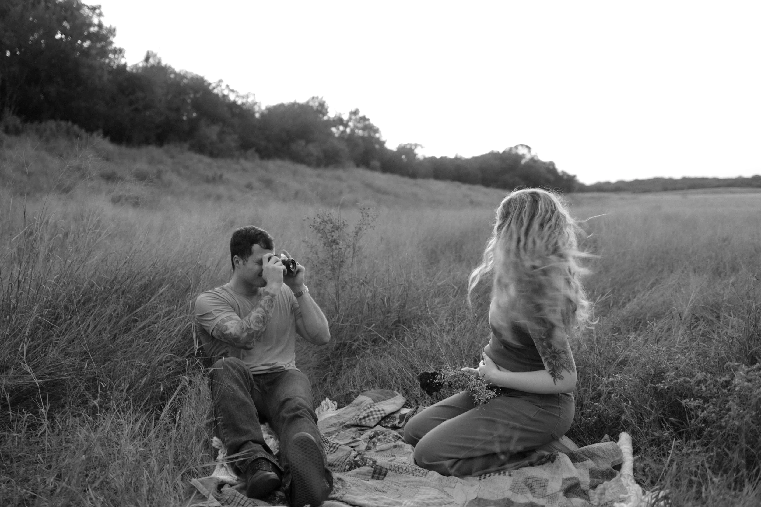 A man taking a photograph of a woman sitting on a blanket in a grassy field during the daytime.