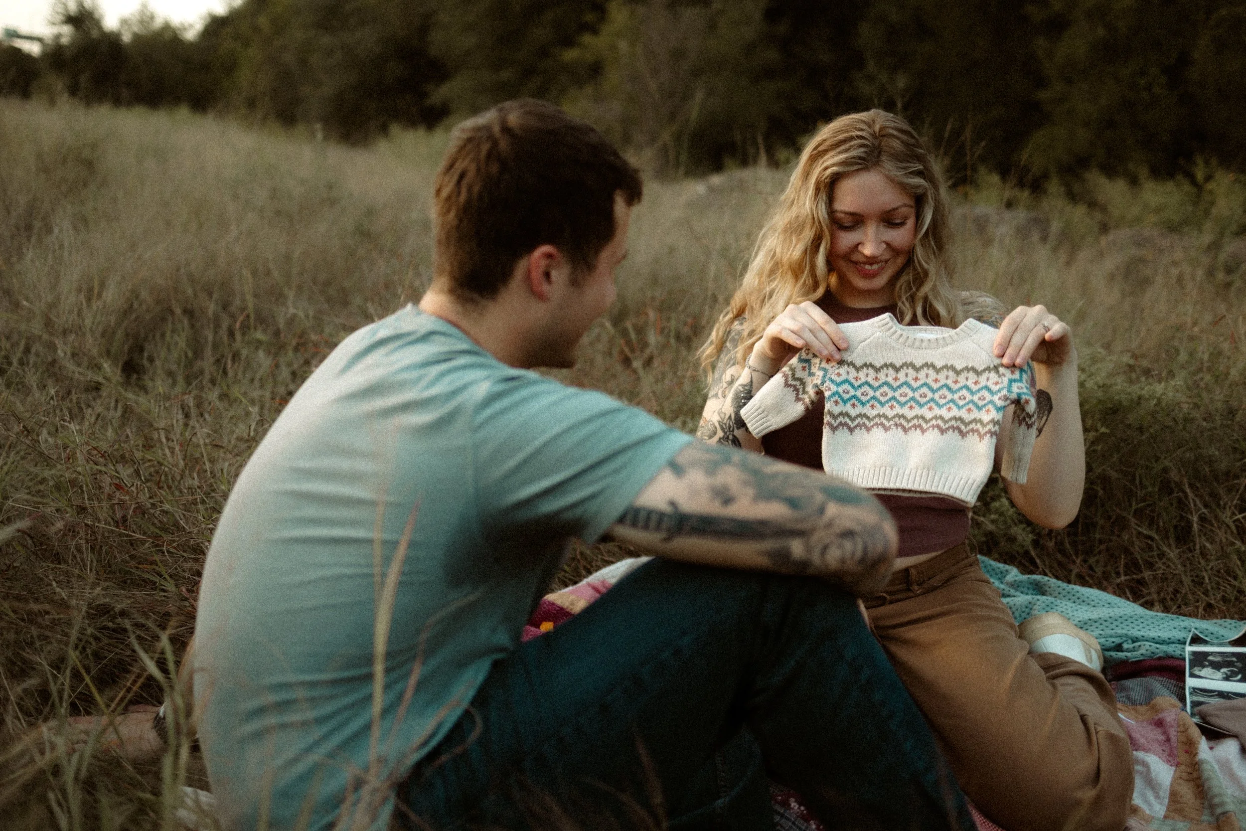 A man and woman sitting in a field, the woman holding a small sweater with a Nordic pattern, smiling and looking at it, while the man looks on.