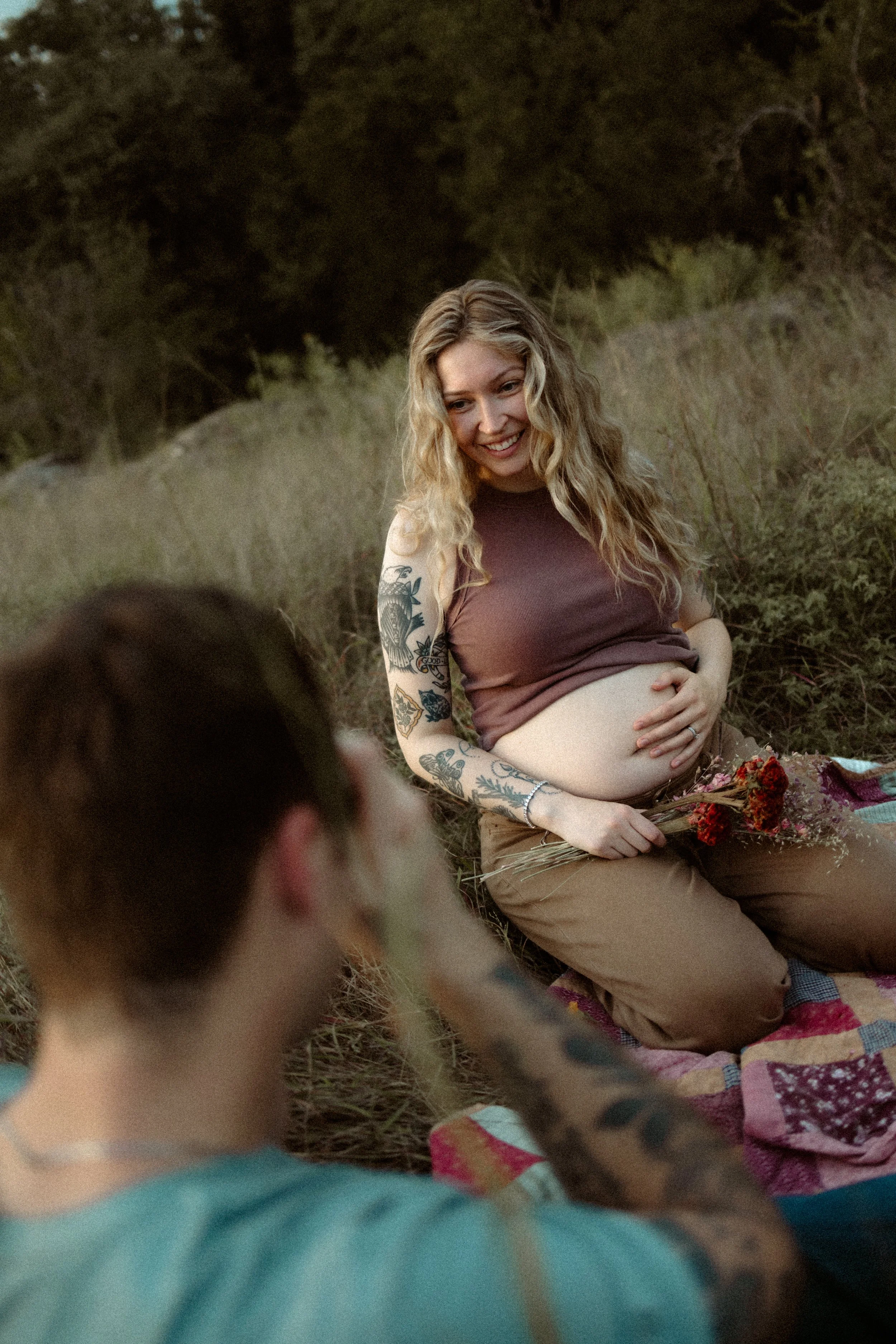 A pregnant woman with tattoos sitting outdoors on a blanket, holding a bouquet of flowers, smiling and looking at a person in the foreground, in a natural setting with trees and grass.