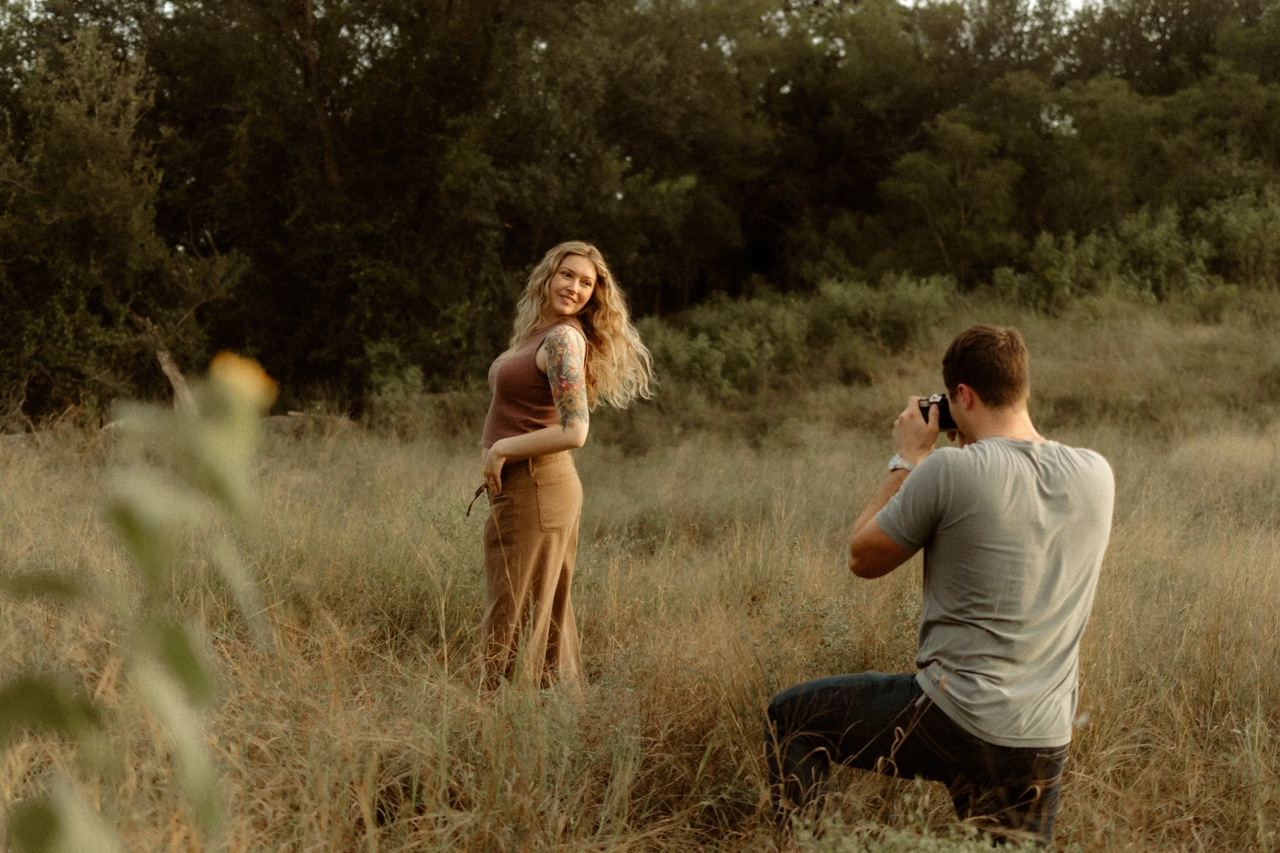 A woman with wavy blonde hair and tattoos posing outdoors while a man kneels taking her photo in a grassy field with trees in the background.