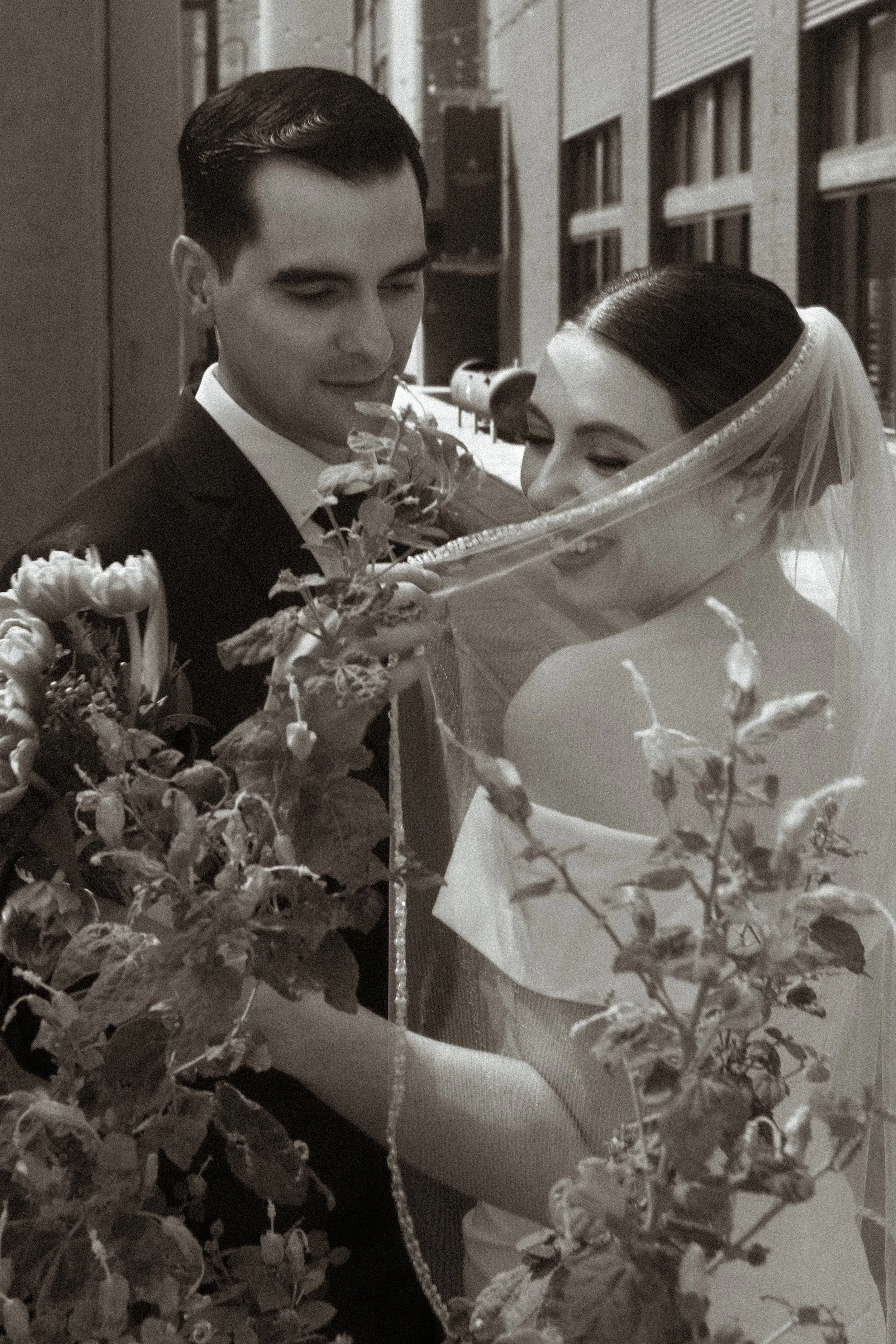 A black-and-white photo of a bride and groom on their wedding day. The groom is in a suit, and the bride is wearing a veil and holding a bouquet of flowers, smiling as they look at each other.