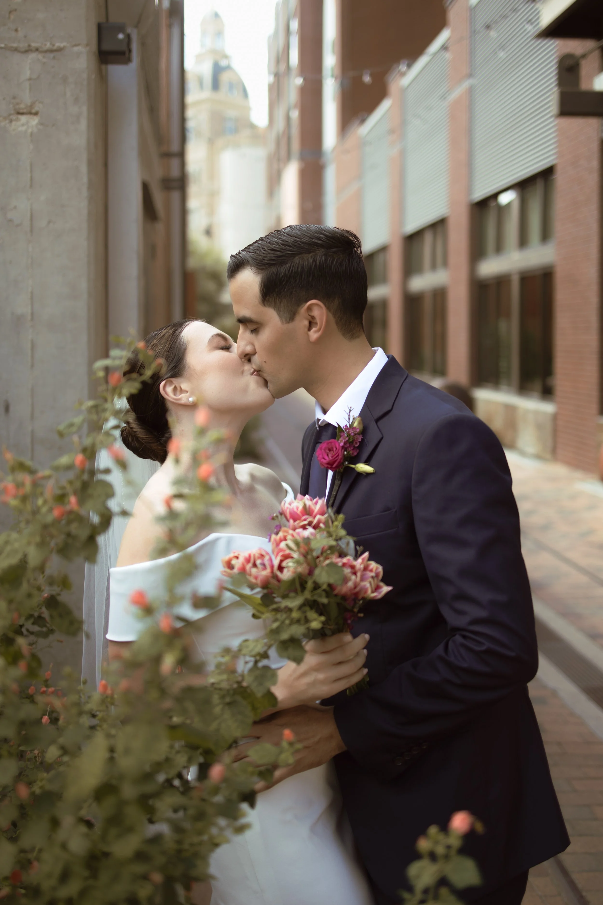 A bride and groom sharing a kiss. The bride holds a bouquet of pink flowers, and they stand in an alley with brick and concrete buildings in the background.