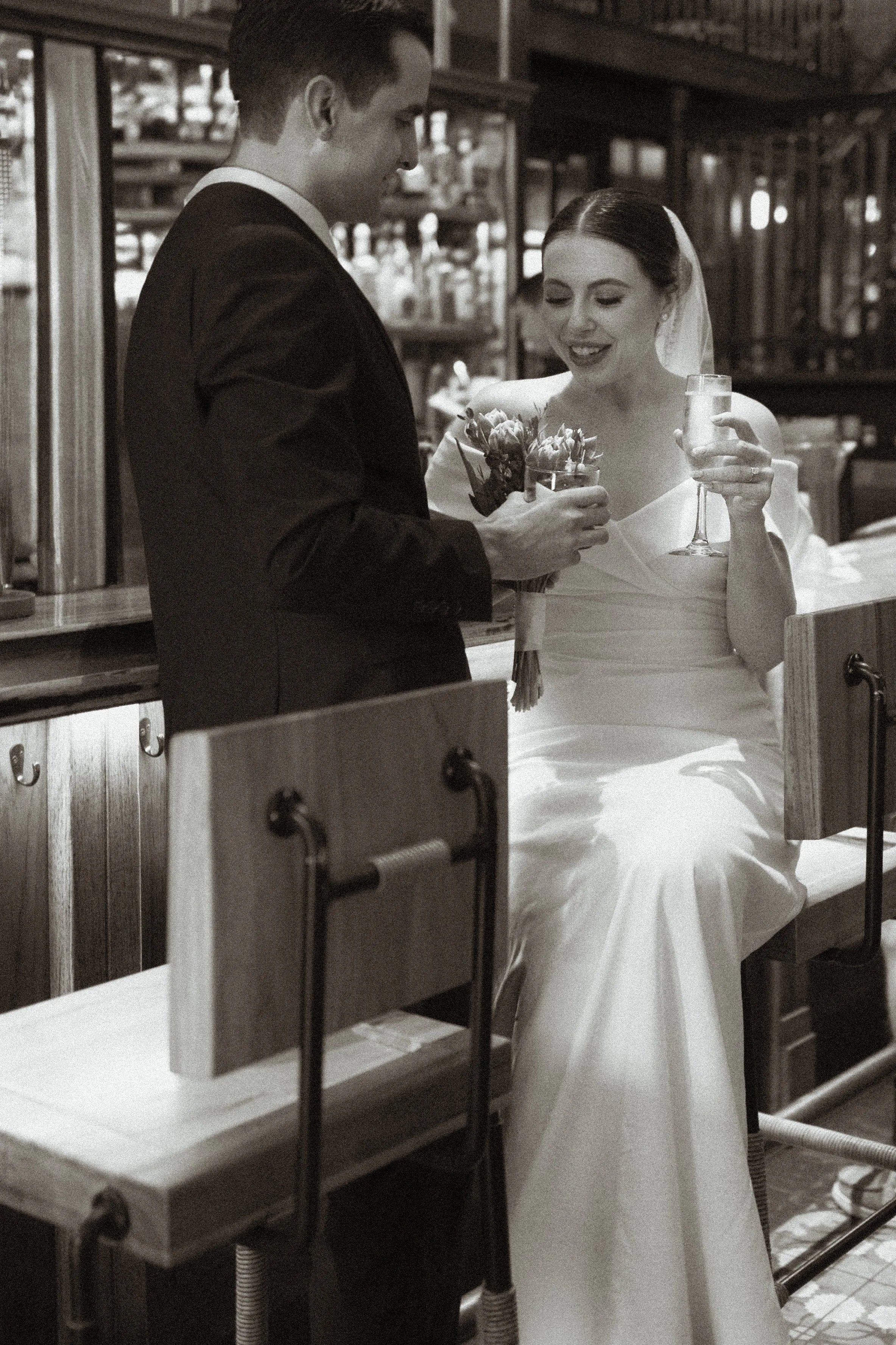 A black and white photo of a bride and groom at a bar, exchanging drinks and flowers, with a rustic setting in the background.