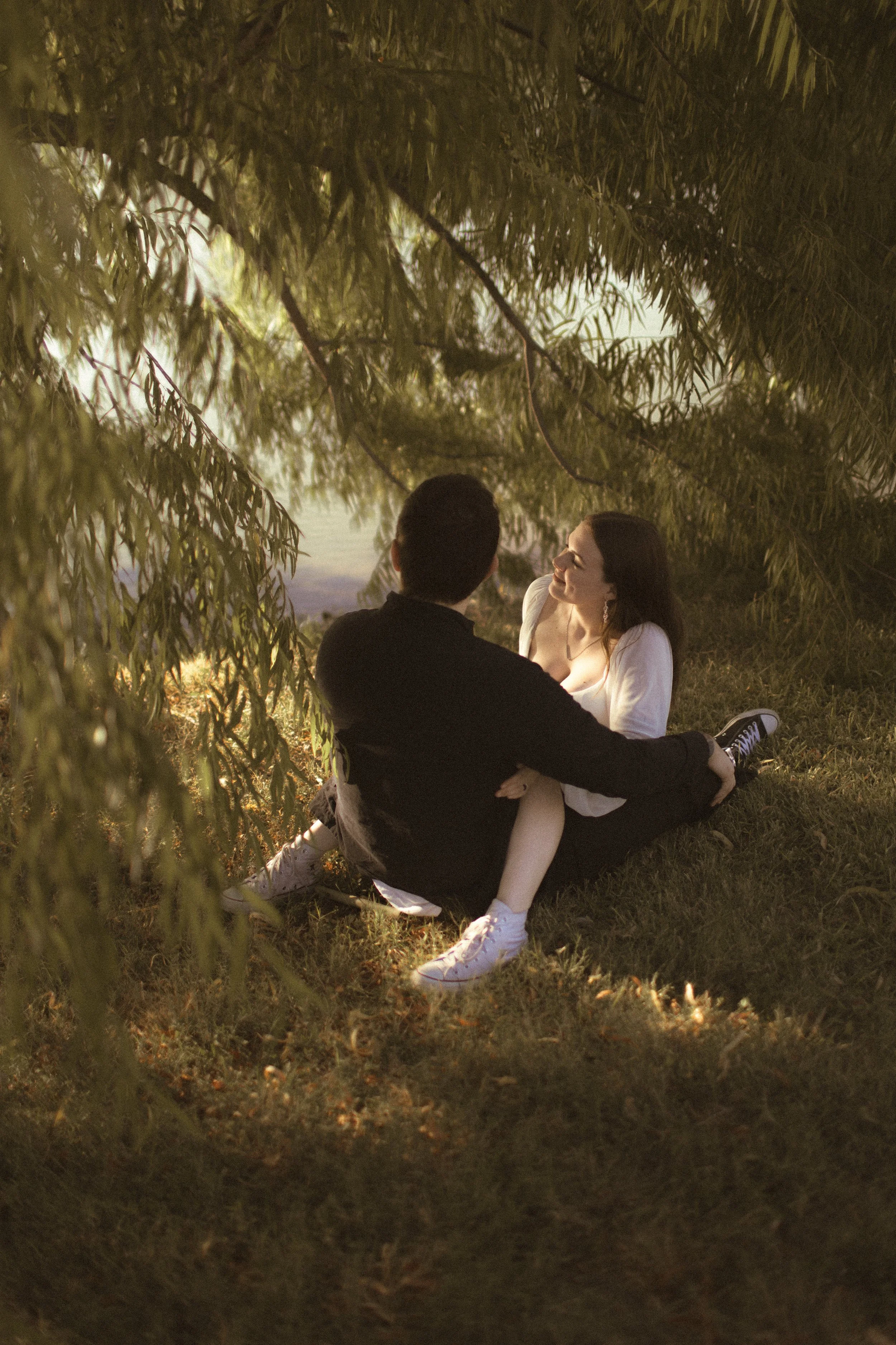 A couple sitting on the grass under a tree, with the woman smiling and looking into the man's face.