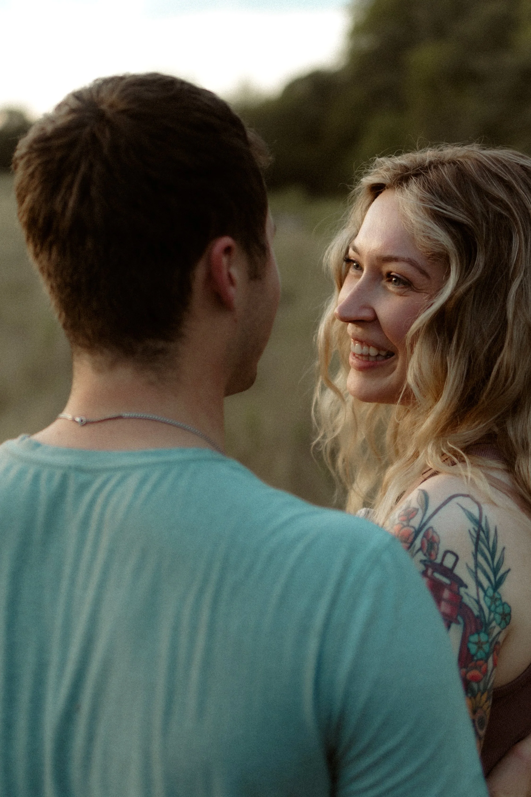 A young couple standing closely outdoors, the woman smiling and looking at the man, with a blurred natural background.