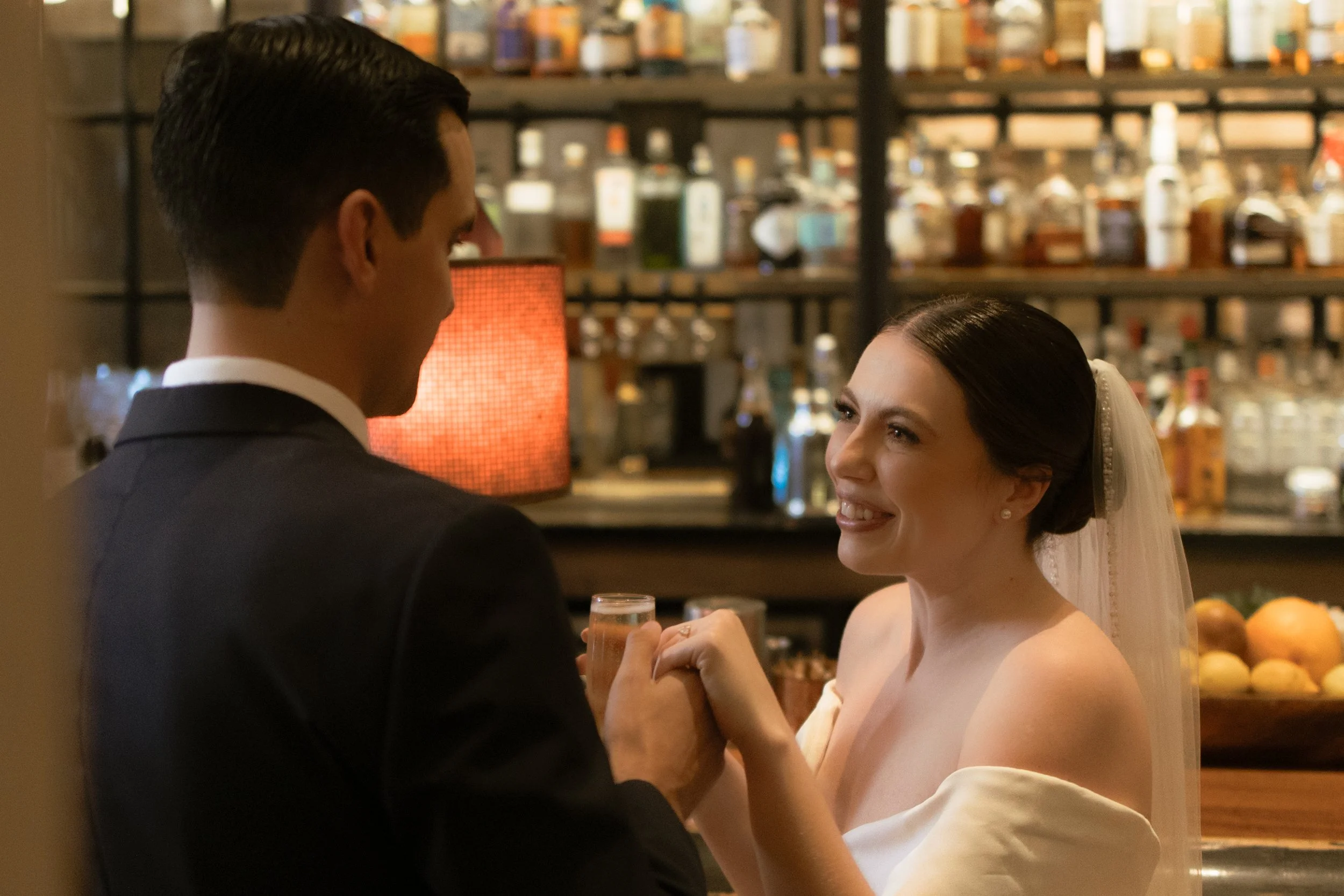 A bride and groom share a moment at their wedding reception, holding hands and smiling at each other, with a bar in the background.