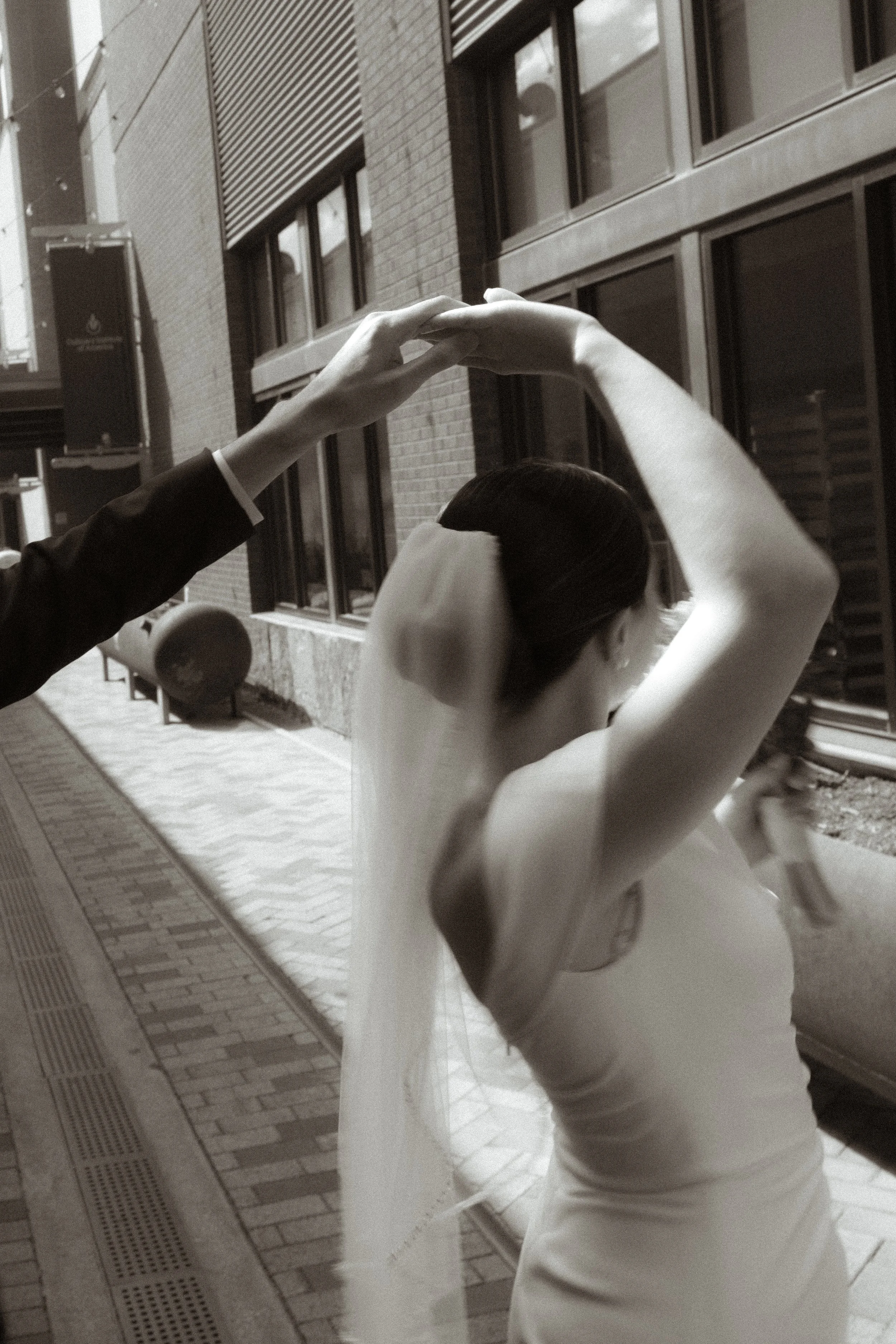 A black and white photo of a woman in a wedding dress with a veil, posing outdoors with her arms raised above her head, while someone else holds her hand.