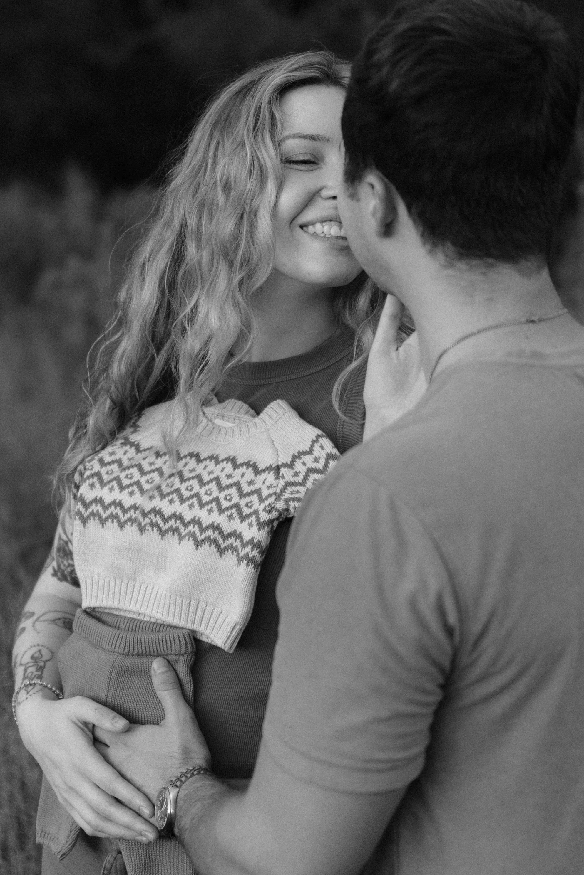 A woman and a man are close together outdoors, with the woman smiling and her eyes closed, and the man touching her chin.