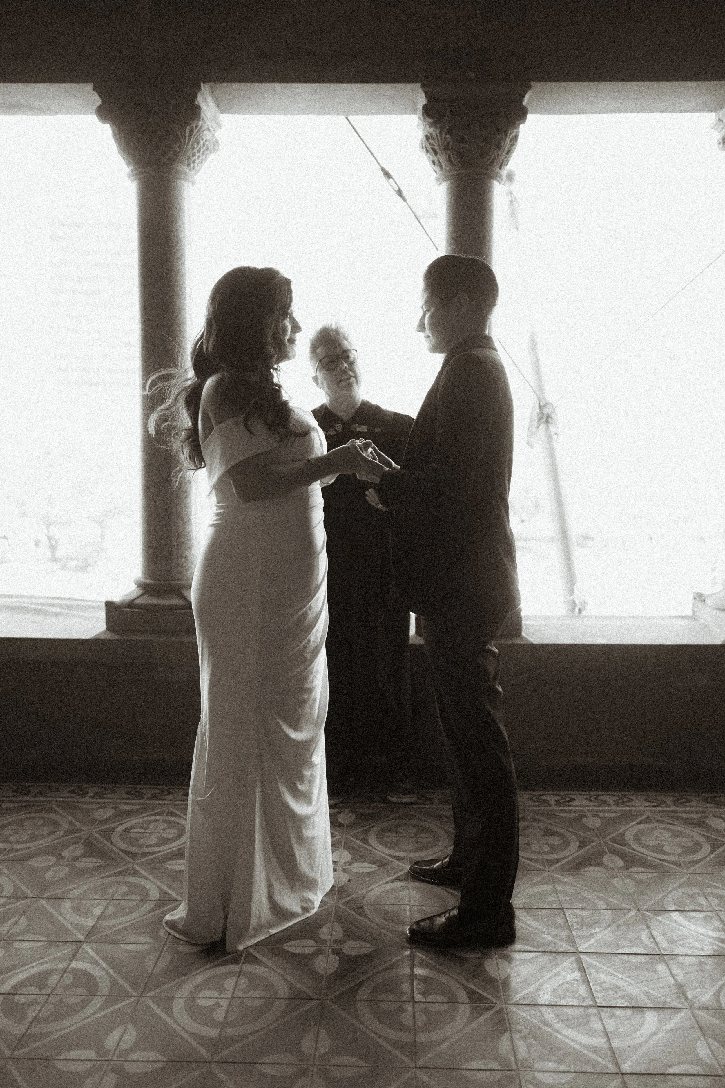 A black and white photo of a wedding ceremony with a bride and groom holding hands and standing before an officiant in front of large windows with decorative columns.