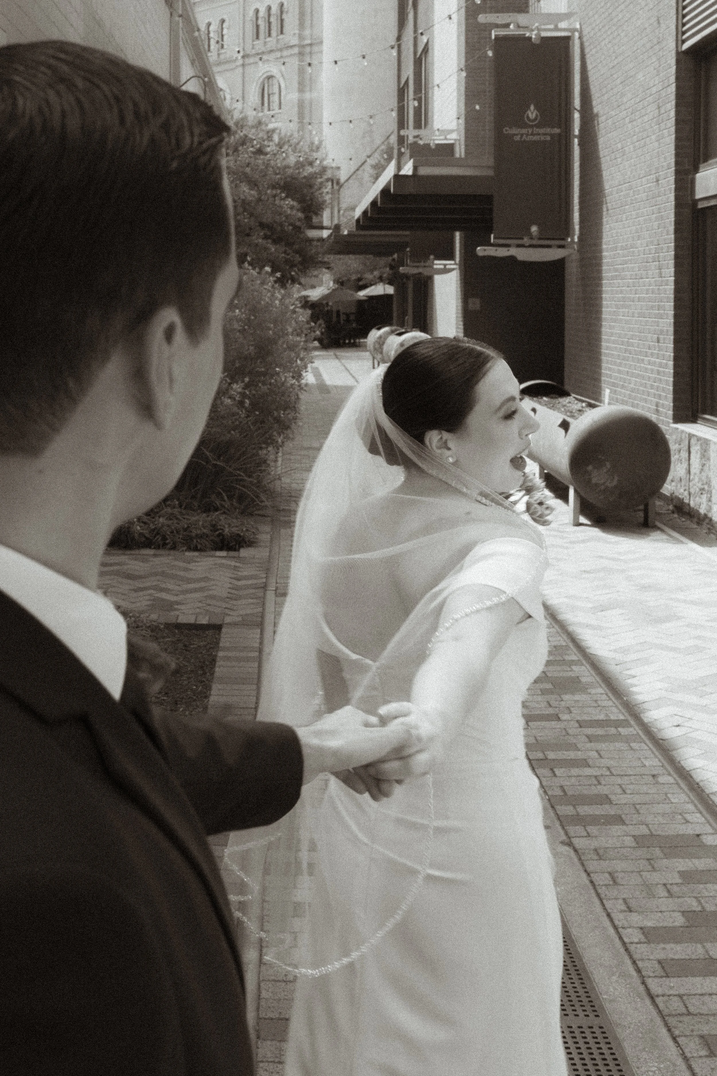 A bride and groom holding hands outdoors, with the bride smiling and wearing a wedding dress and veil, and the groom in a suit, on a city street.