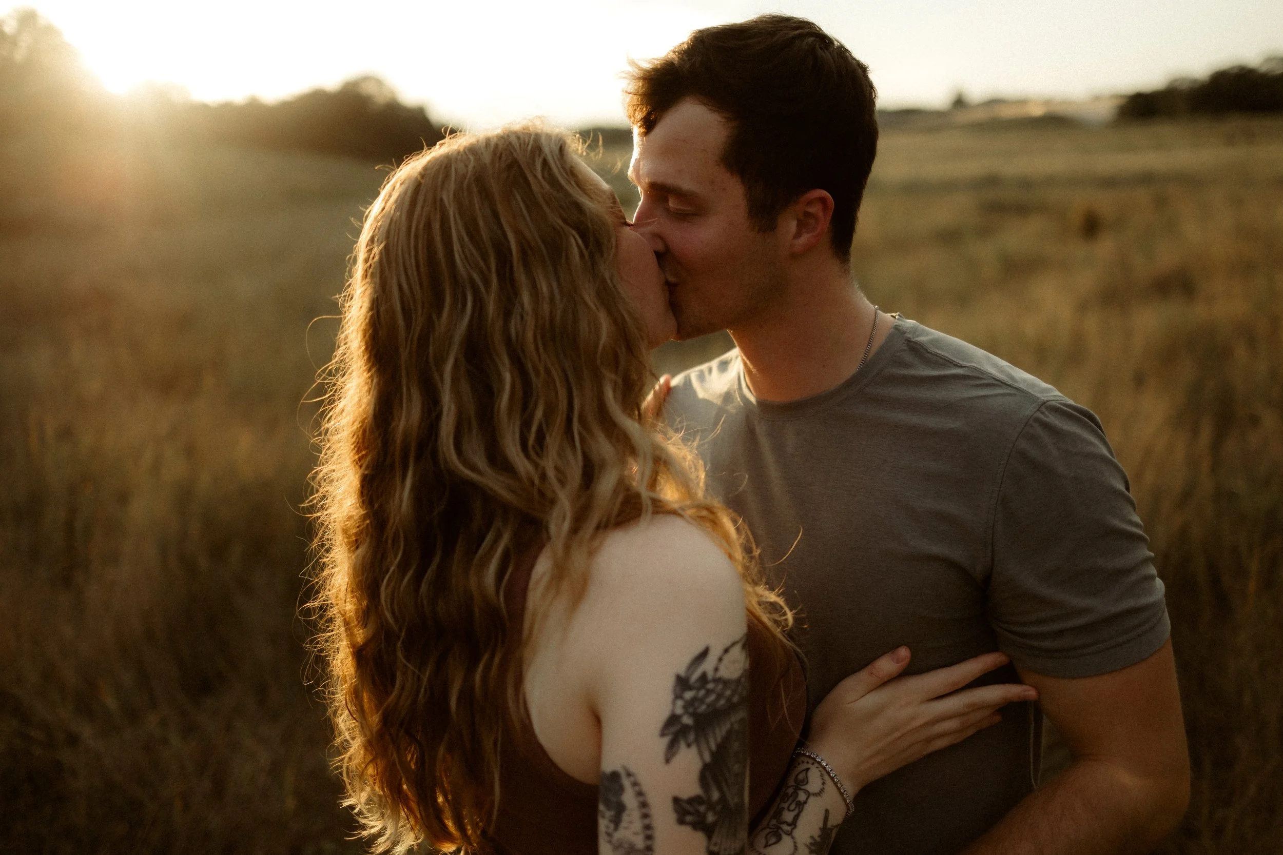 Couple kissing in a field during sunset, with the woman having long curly hair and tattoos on her arm, and the man wearing a gray T-shirt.