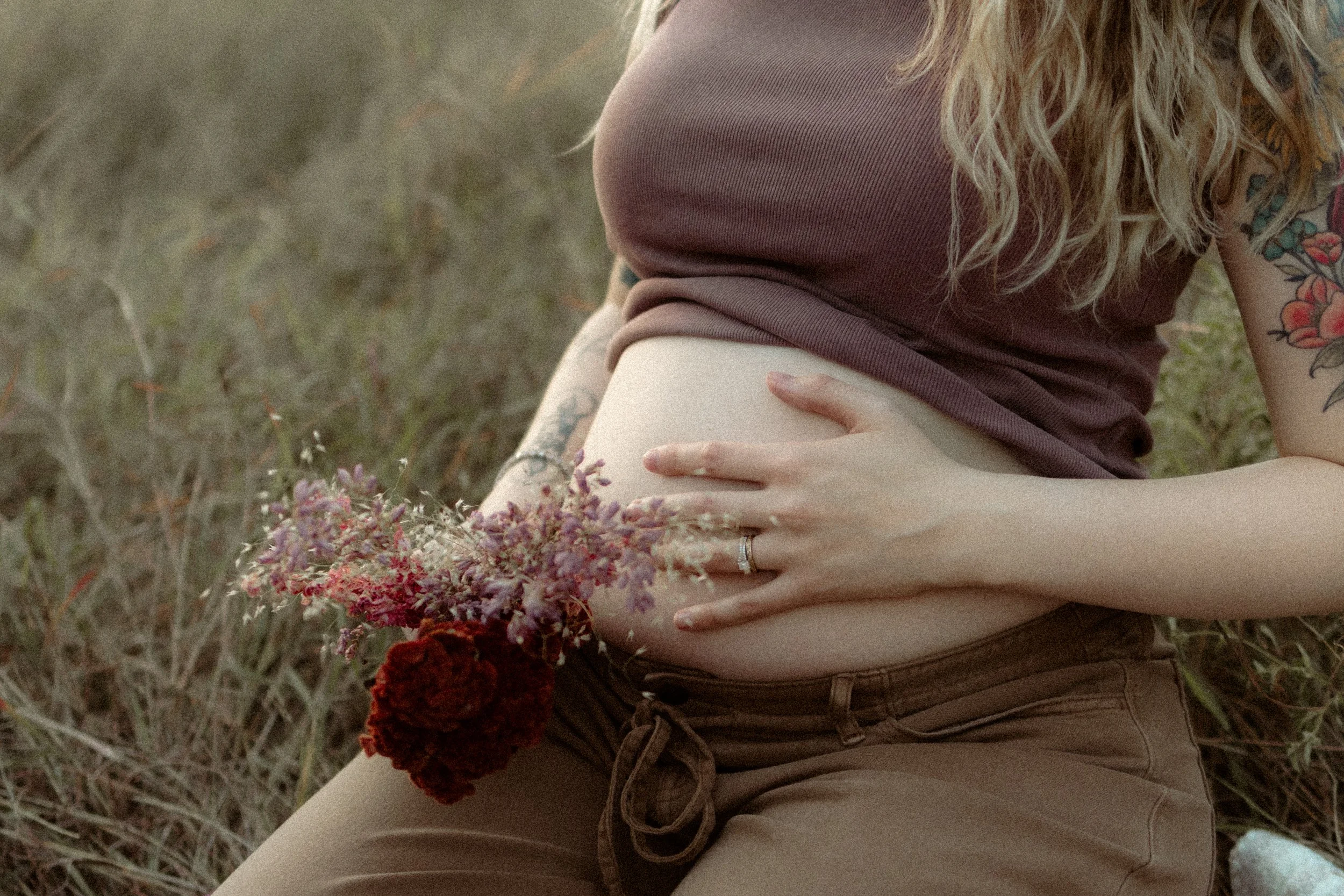 A pregnant woman is sitting in a grassy field, holding her belly with one hand and a bouquet of flowers with the other