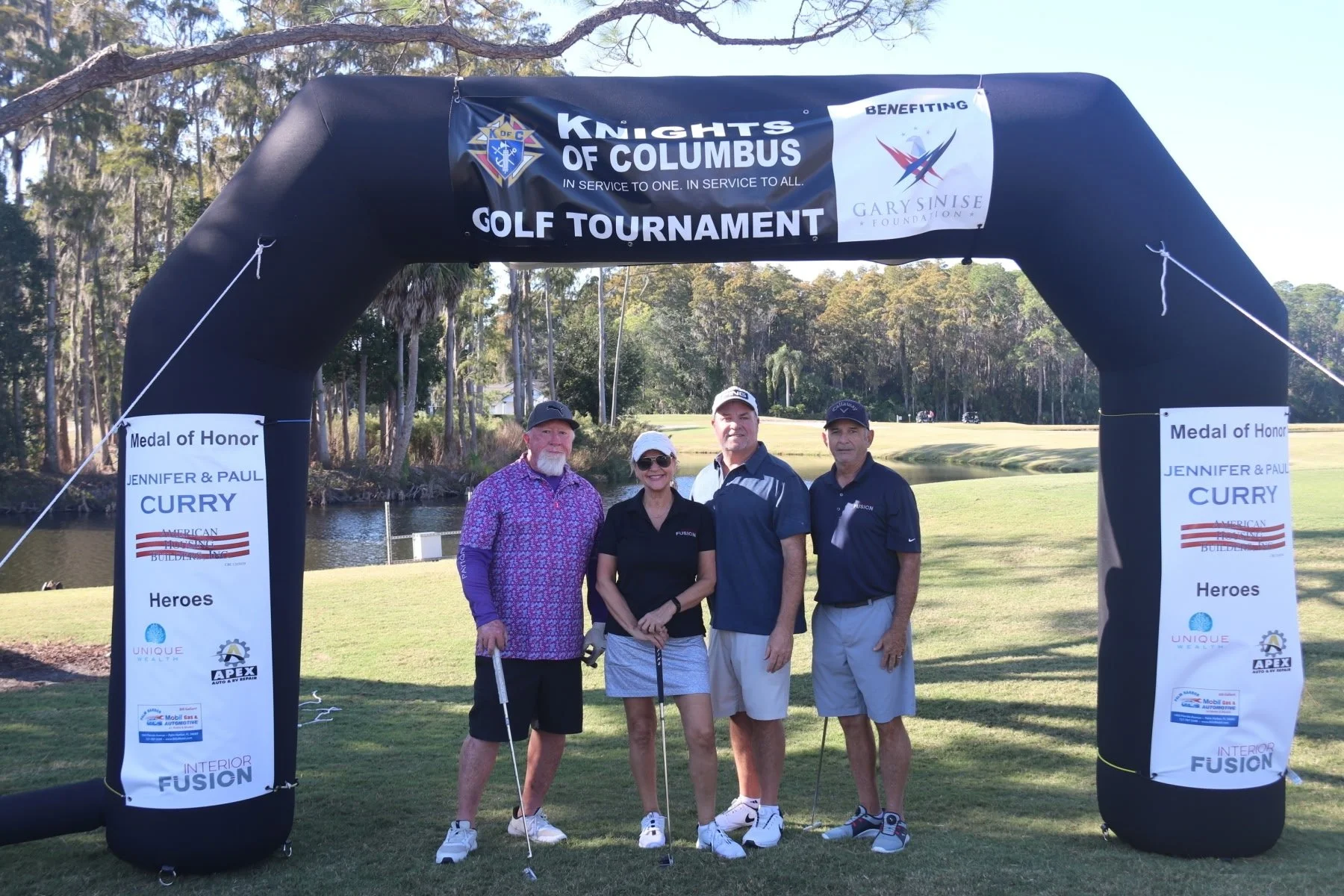 Four golfers posing under Knights of Columbus Golf Tournament inflatable archway benefiting Gary Sinise Foundation with Medal of Honor sponsor banners for Jennifer and Paul Curry.
