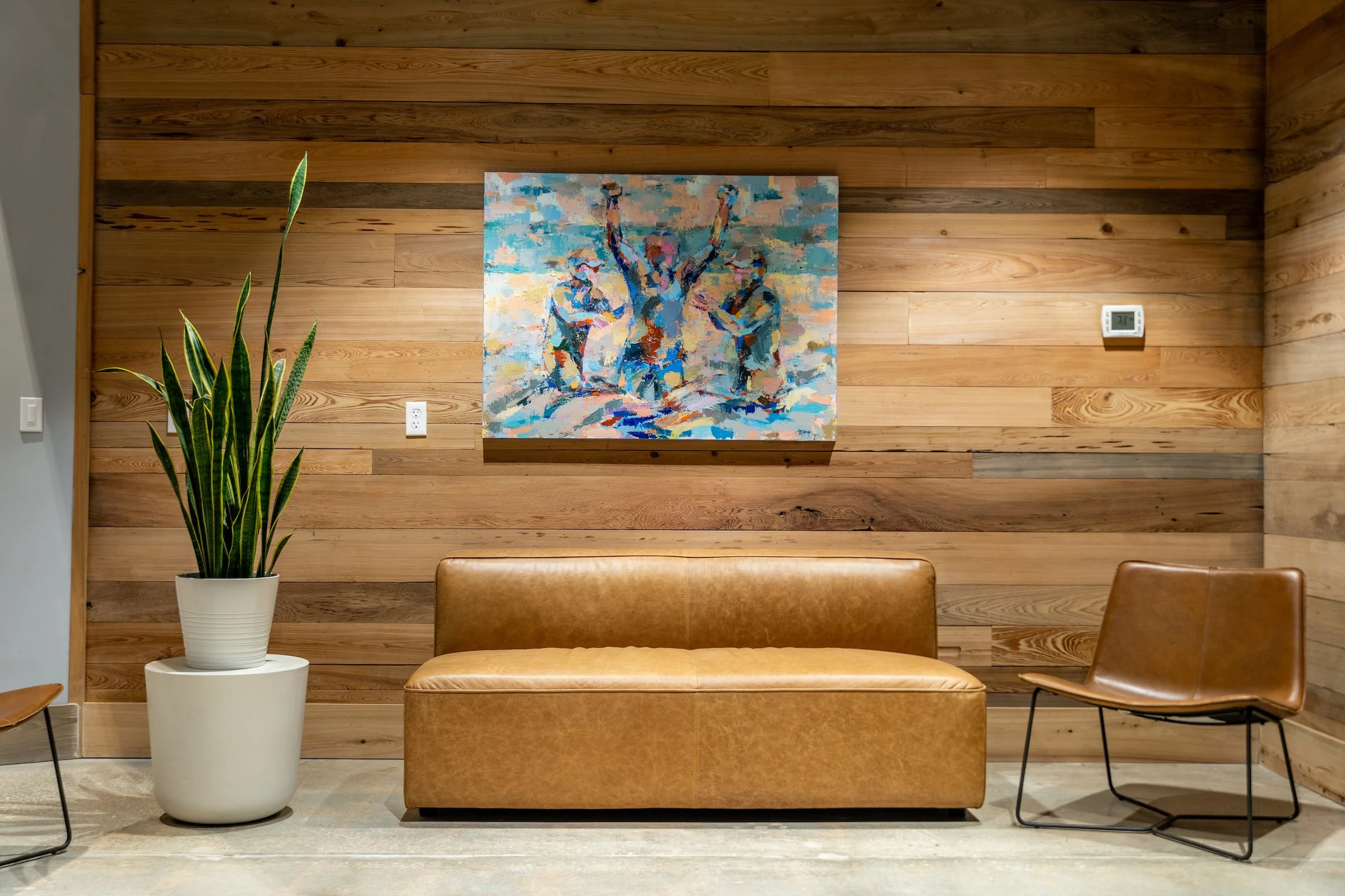 Office lounge area with reclaimed wood accent wall, leather sofa and chair, snake plant, and colorful abstract artwork.