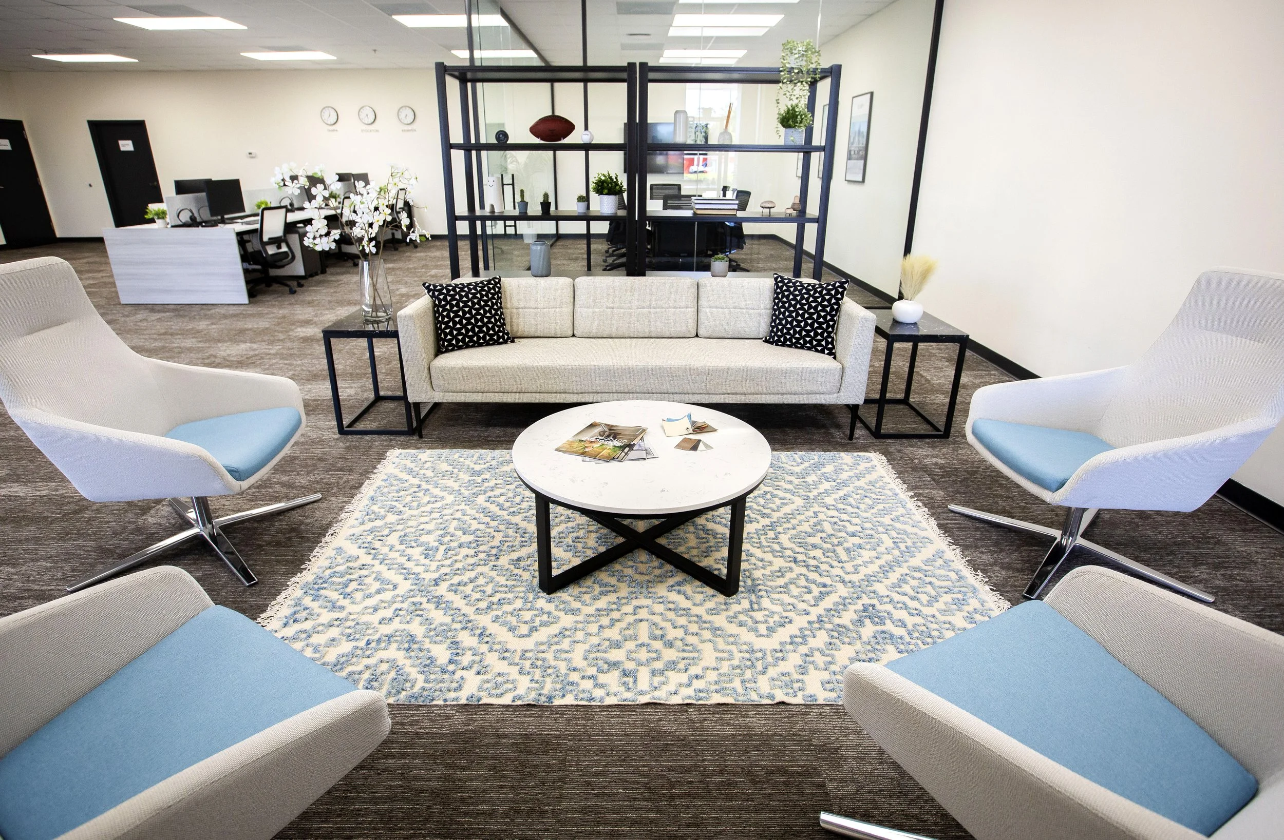 Modern office lobby with beige sofa, white accent chairs with blue cushions, and contemporary shelving divider.