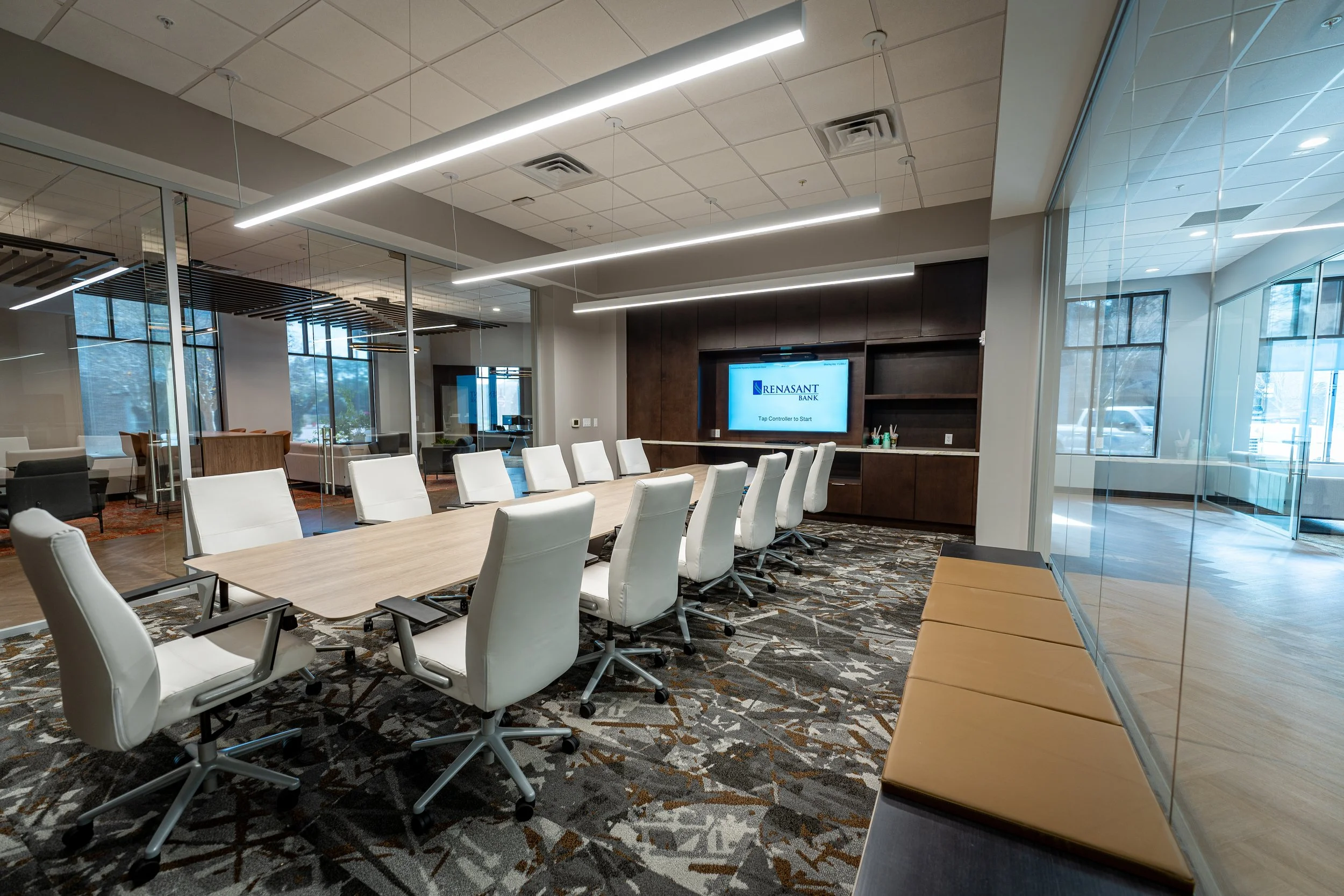 Modern Renasant Bank conference room with wooden table, white chairs, dark wood cabinetry, mounted display screen, and glass-enclosed offices.