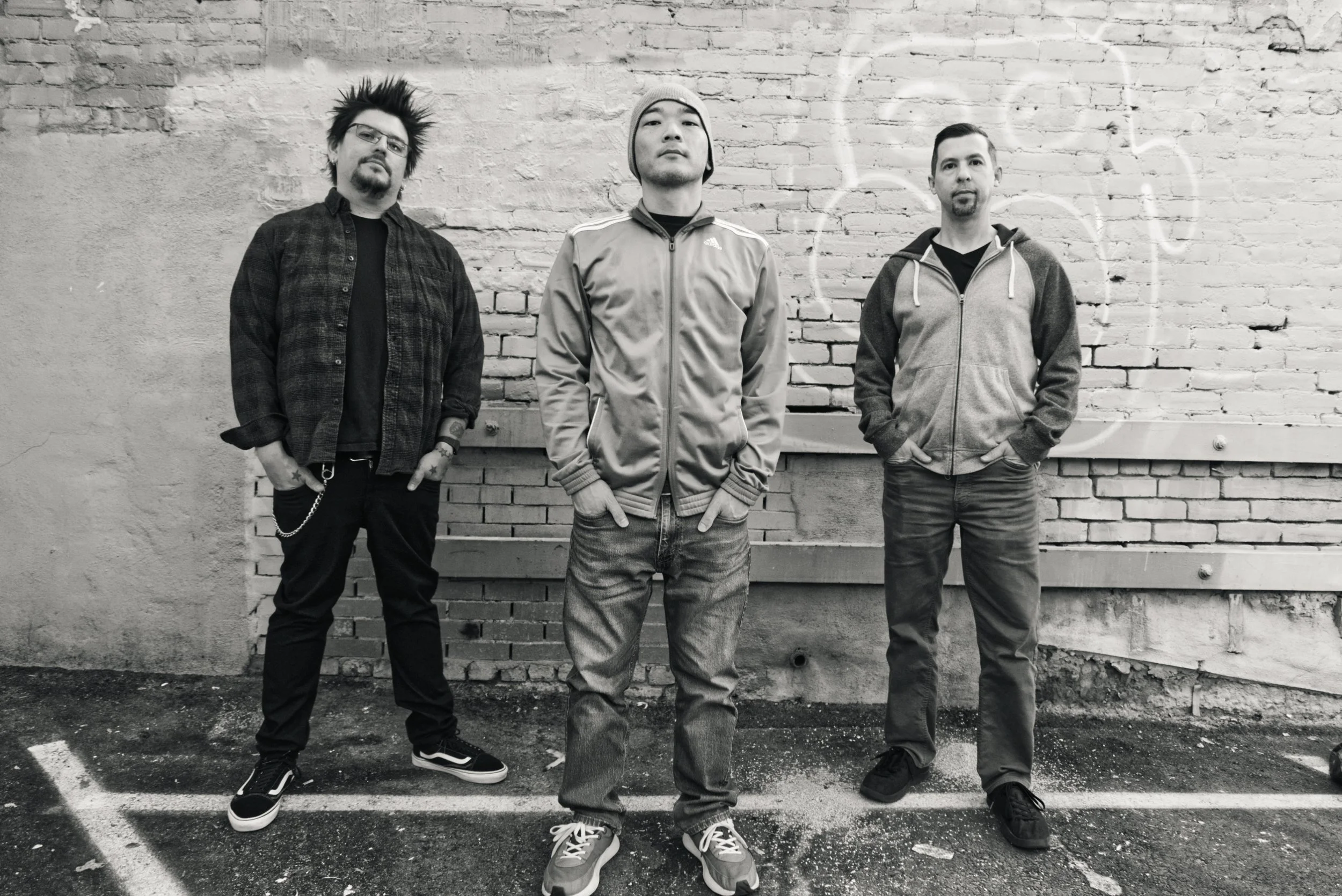 Three band members standing in front of a brick wall with faint graffiti, dressed casually in jackets and jeans in a parking lot.
