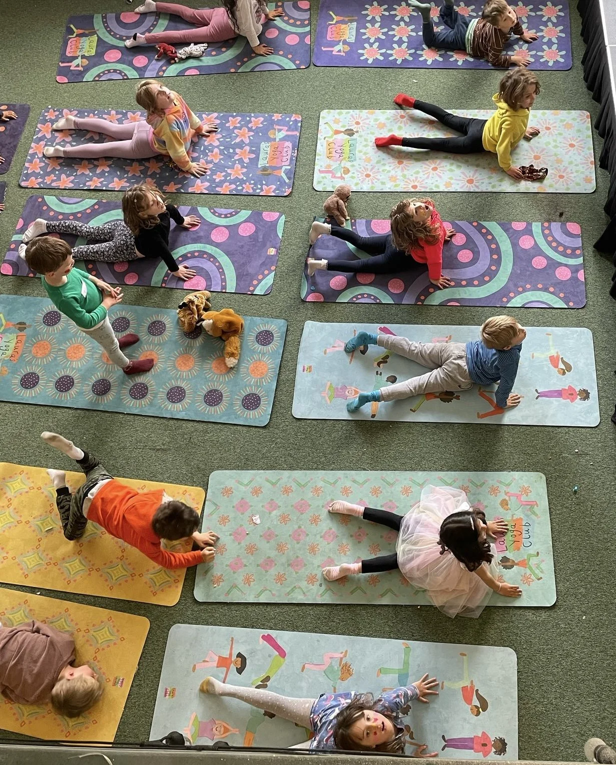 Children are participating in a yoga class, lying on colorful yoga mats on a green carpeted floor, with some kids and stuffed animals nearby, engaging in stretching and balancing exercises.