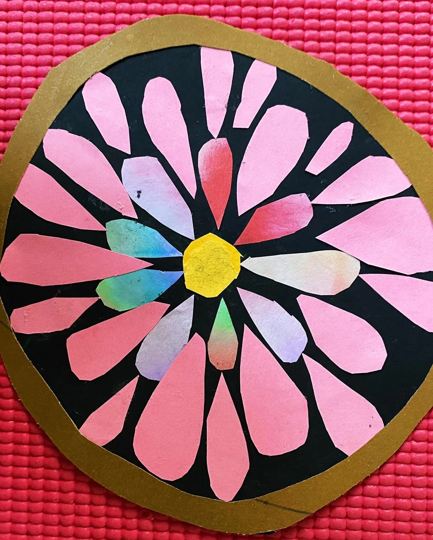 Decorative paper craft resembling a colorful flower with pink petals, a yellow center, and a black background, framed with gold border, placed on a textured pink surface.