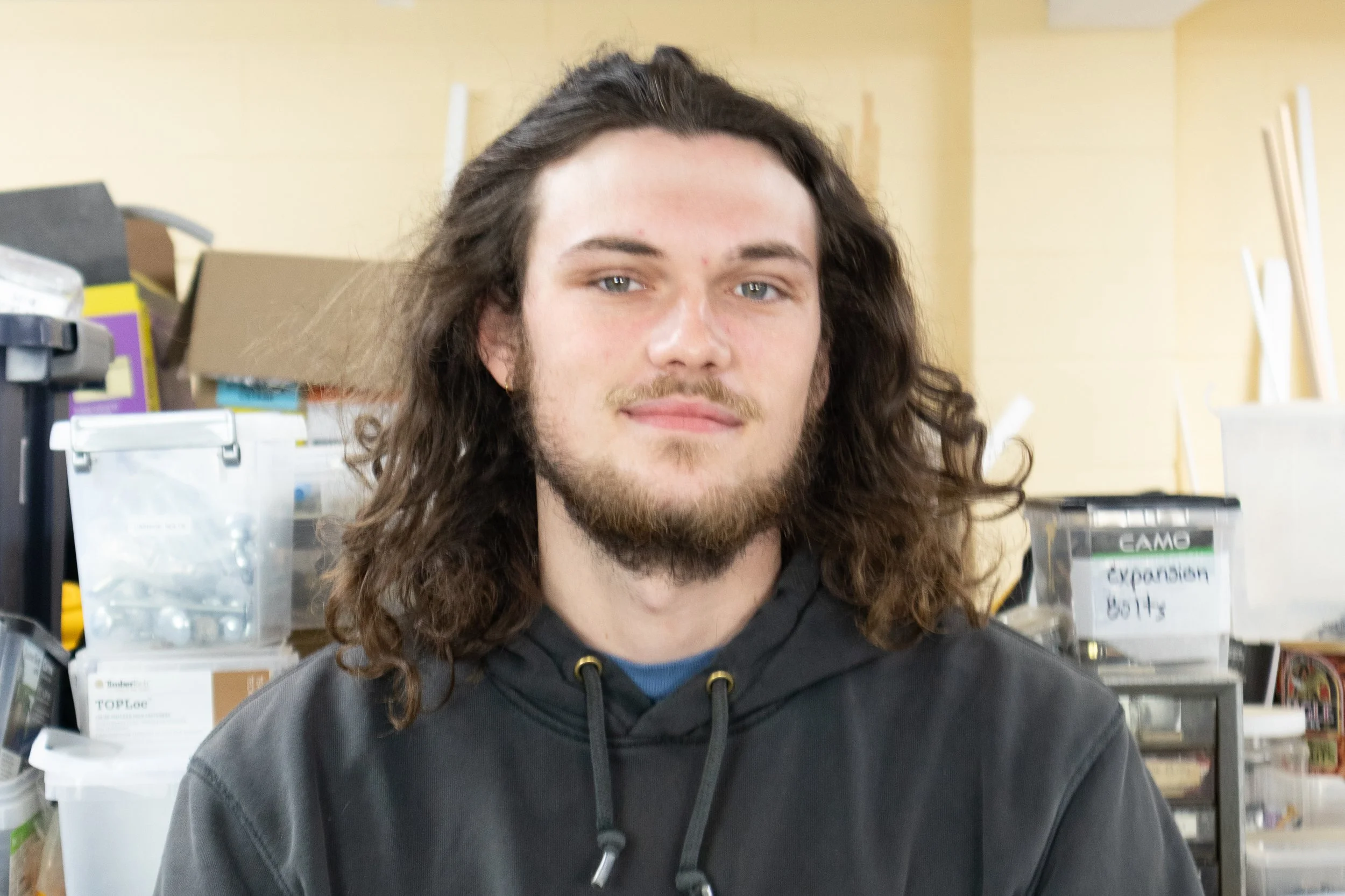 A young man with long, curly brown hair, a light beard, and light-colored eyes, wearing a black hoodie, sitting indoors with shelves and storage boxes in the background.