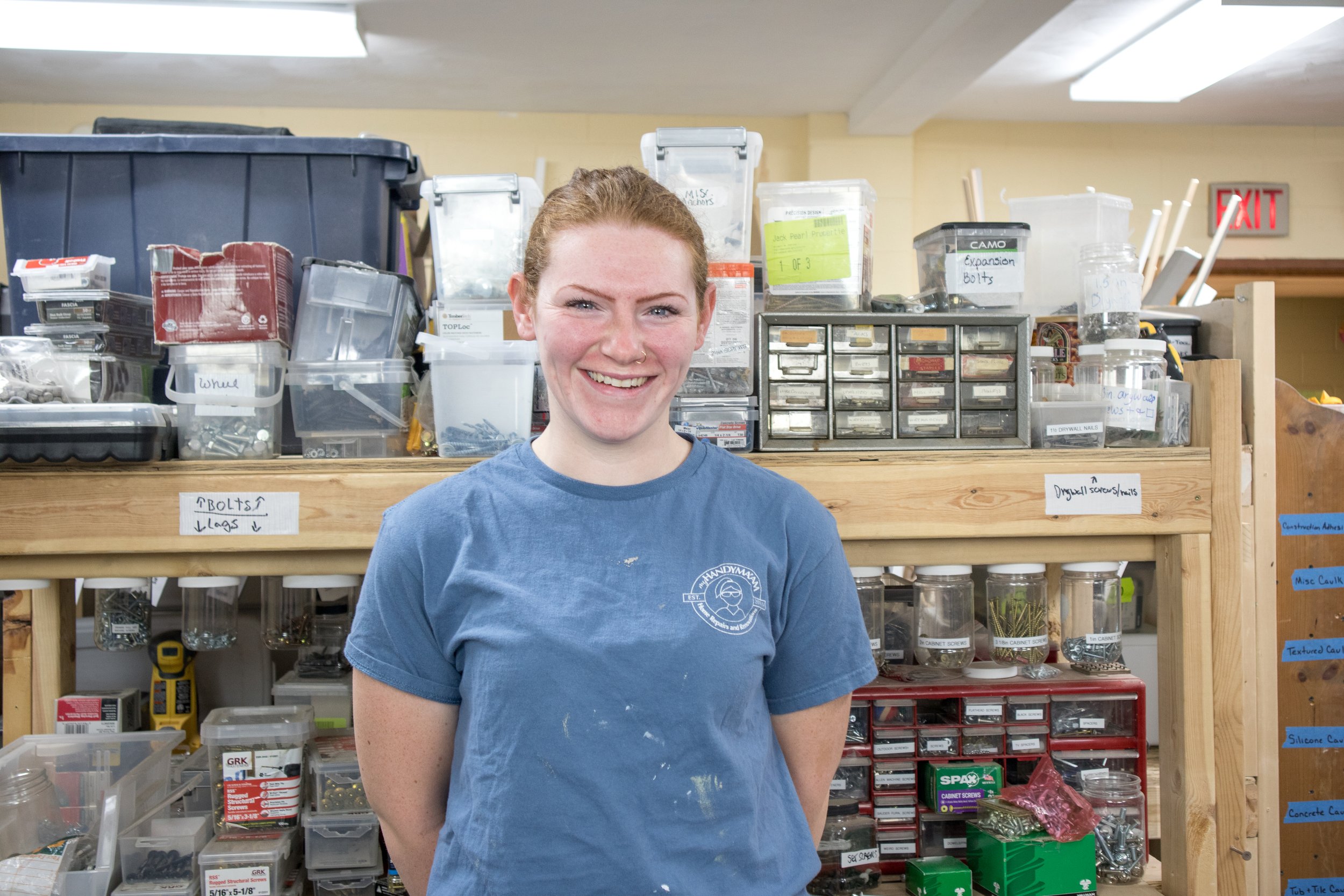 A woman with a blue T-shirt standing in a workshop with organized containers and supplies in the background.